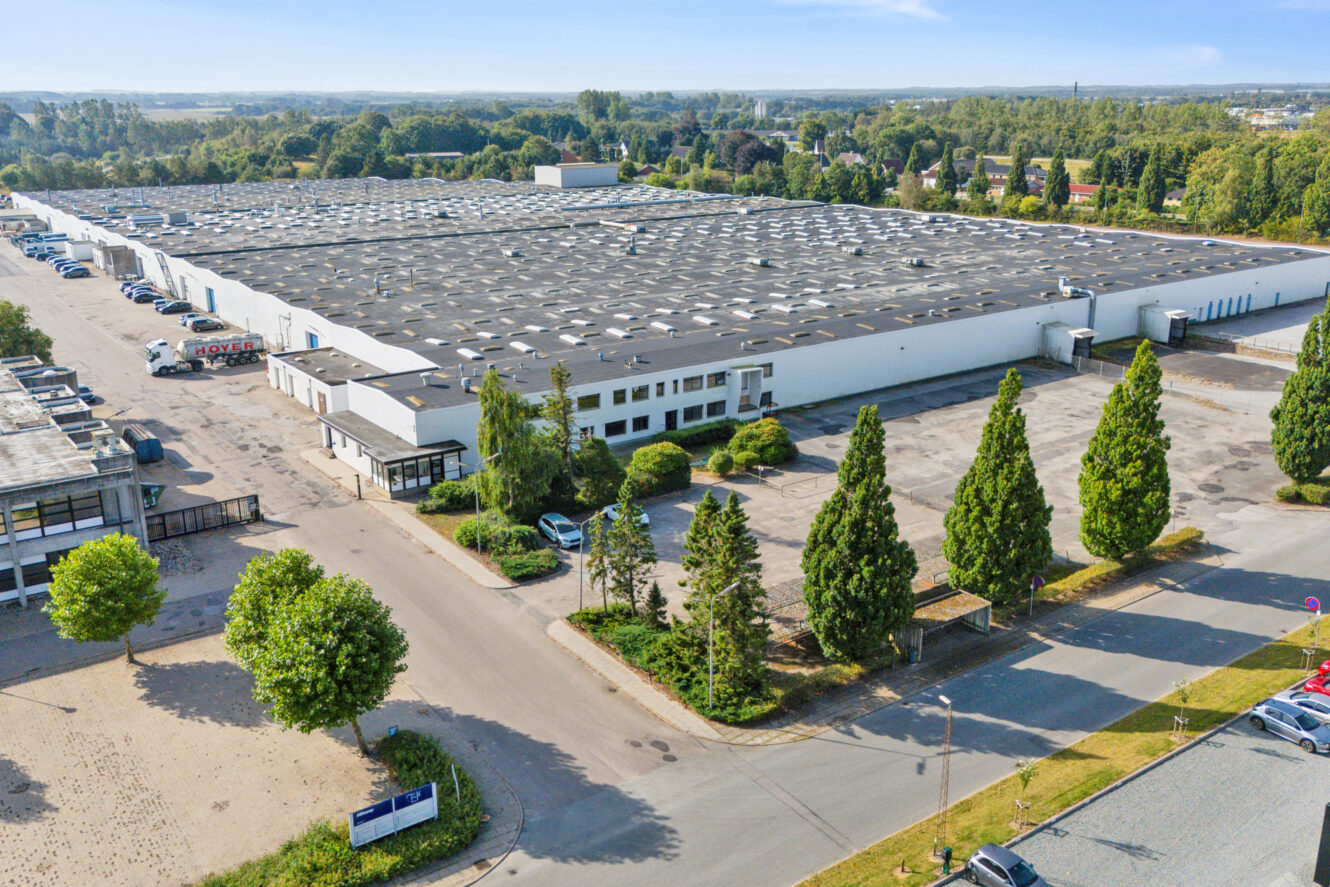 Et stort industrilager med et hvidt ydre, omgivet af træer, parkeringspladser og adskillige lastbiler og biler under en klar blå himmel.