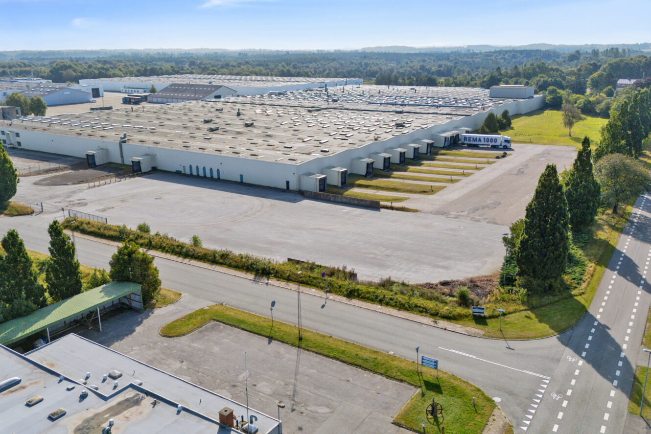 Et stort industrilager med flere ovenlysvinduer og en tom parkeringsplads, omgivet af grønne områder og en vej i forgrunden.