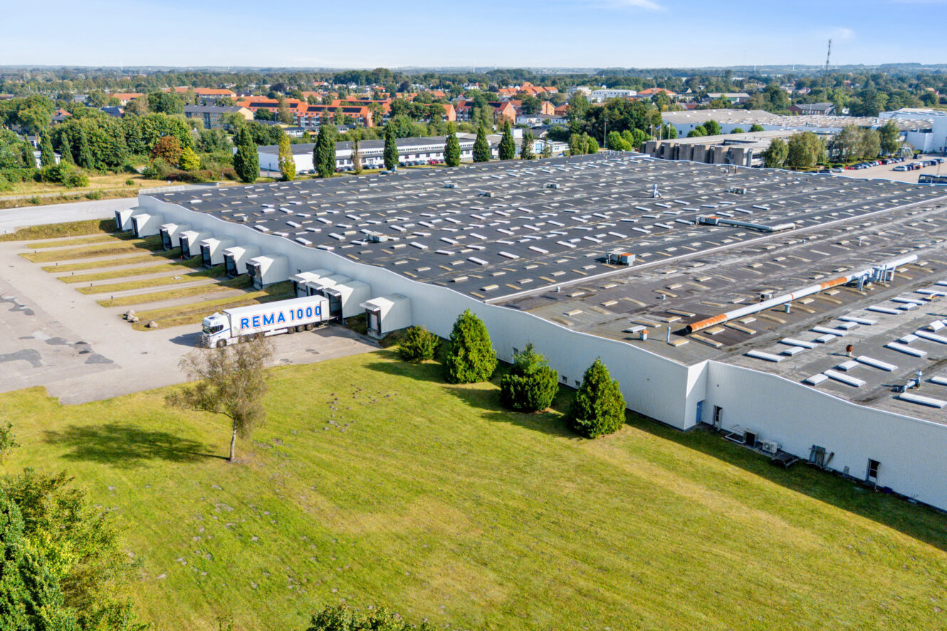 Et stort industrilager med en REMA 1000-lastbil parkeret ved en læsserampe, omgivet af græs, træer og nærliggende bygninger under en klar himmel.