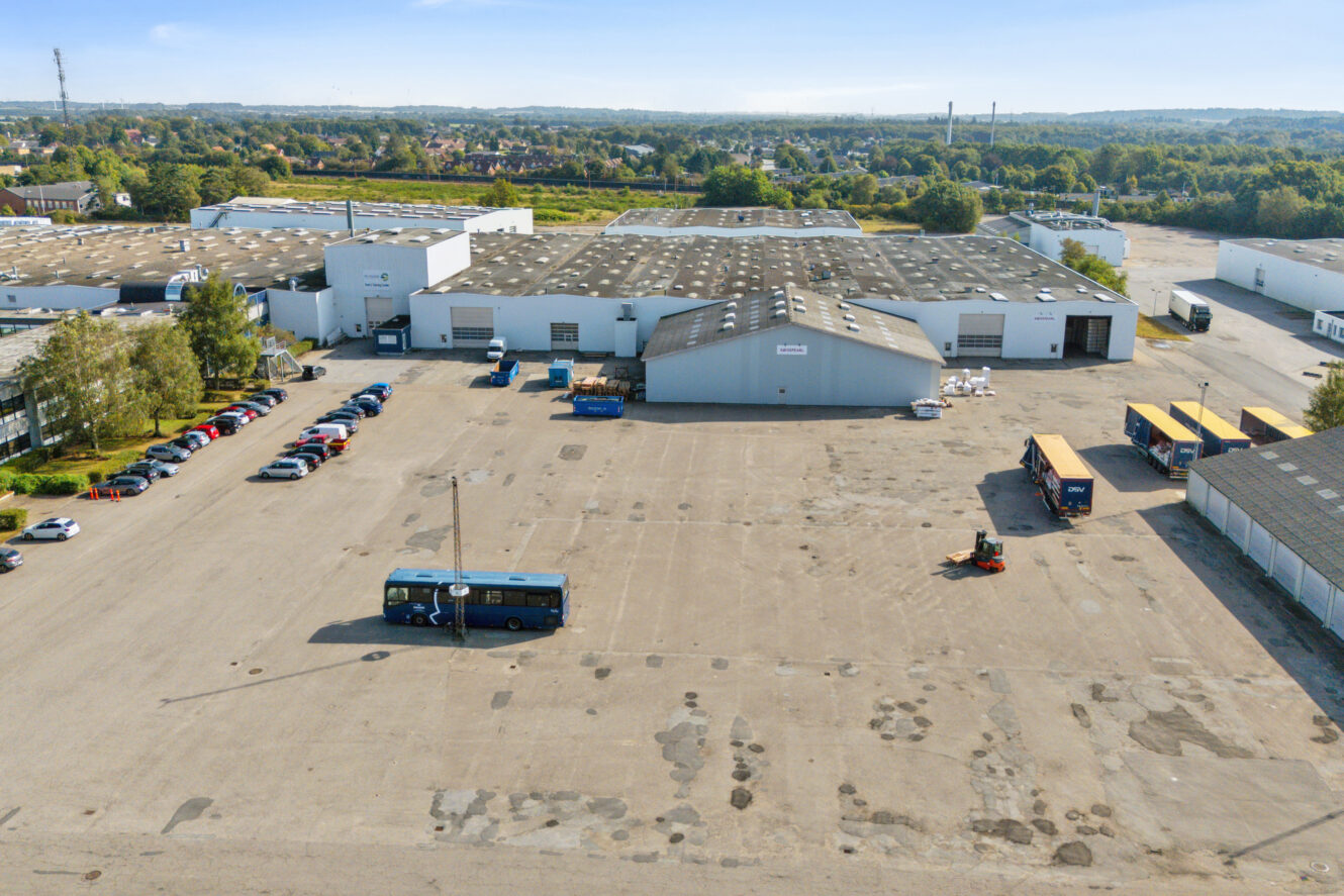 Et stort industrielt lagerkompleks med flere bygninger, parkerede biler, en bus og flere lastbiler på en stor asfalteret plads under en klar himmel.