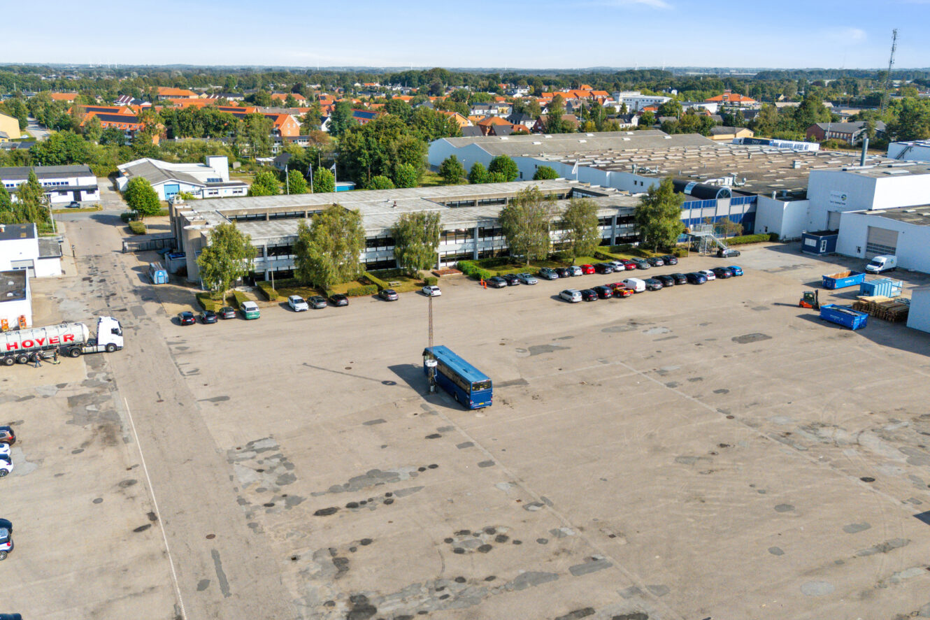 En stor industriparkeringsplads med flere parkerede biler og en blå bus, omgivet af lagerbygninger og træer under en klar himmel.