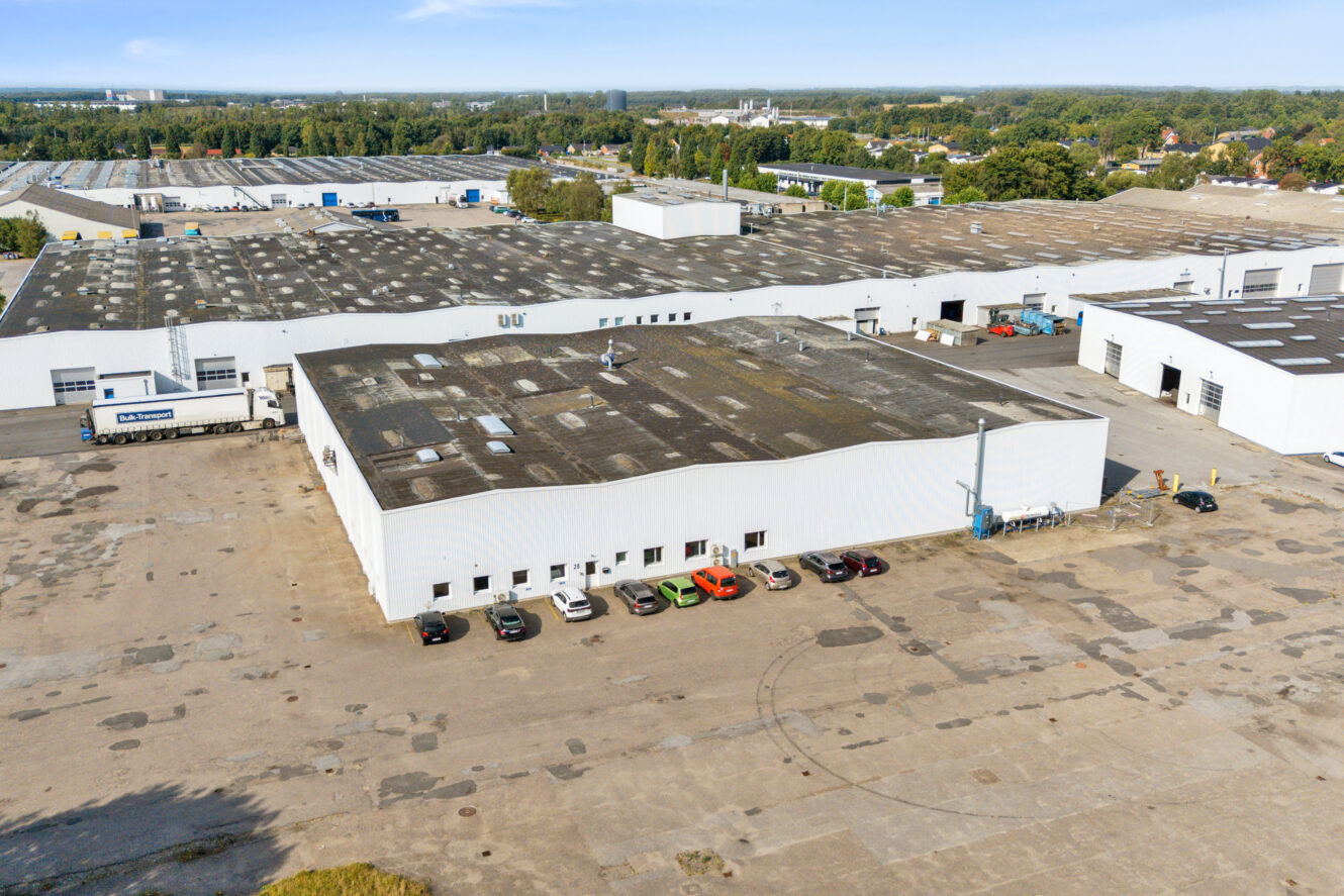 Et stort industrielt lagerkompleks med flere hvide bygninger, parkerede biler, en lastbil og omkringliggende asfalterede områder på en baggrund af træer og fjerne strukturer.