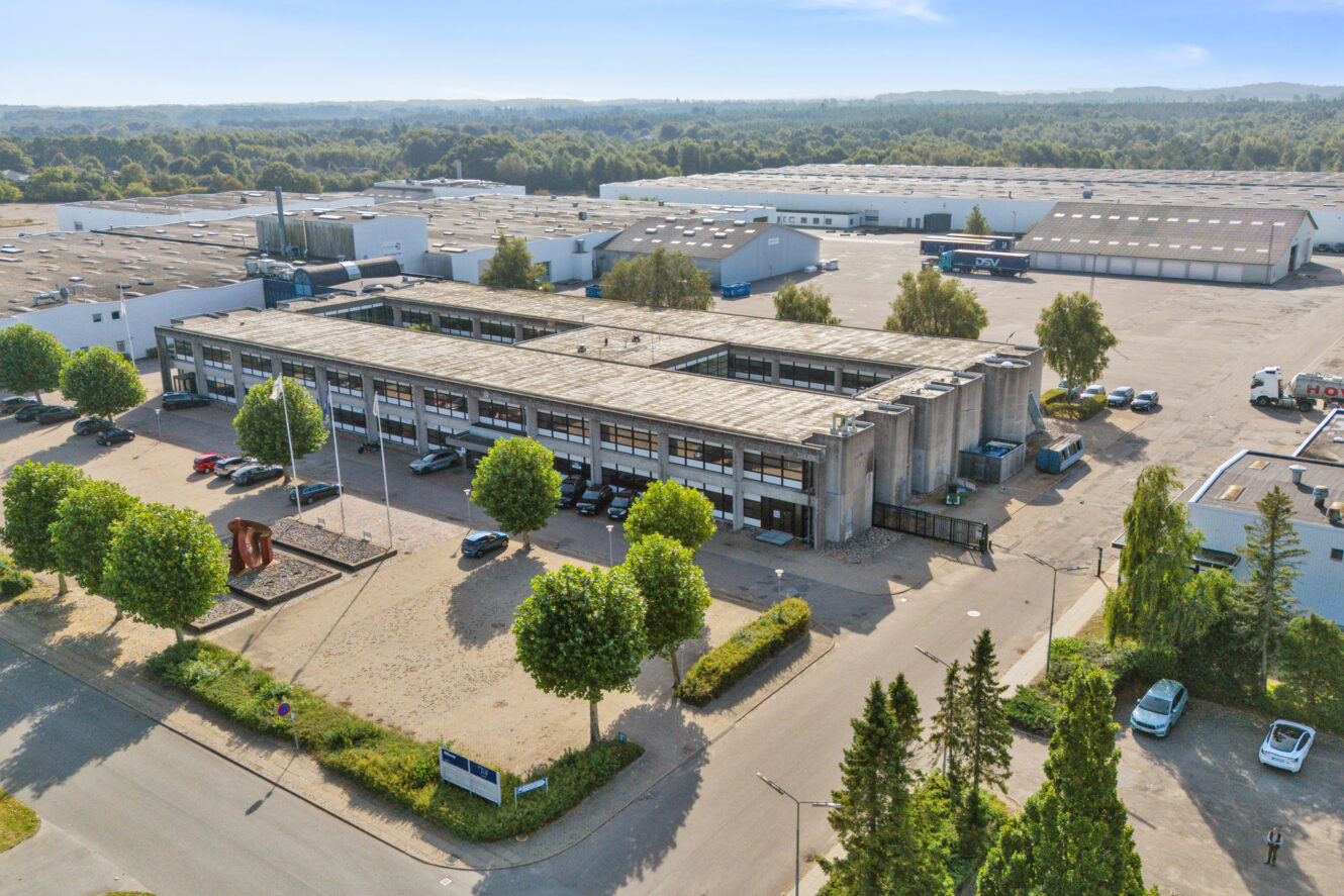 Luftfoto af et industrielt bygningskompleks med parkeringspladser, træer og omkringliggende lagerbygninger under en klar himmel.
