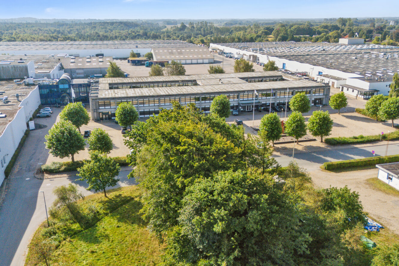 Luftfoto af et industrikompleks med flere store lagerbygninger, parkeringspladser og træbeklædte gader på en klar dag.