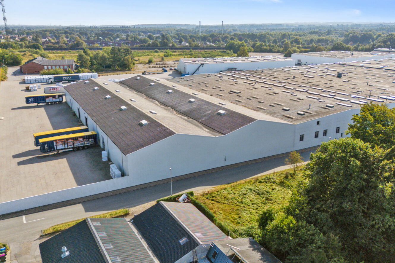 Stort industrilager med læsseramper og lastbiler parkeret udenfor, omgivet af træer og andre bygninger på en klar dag.