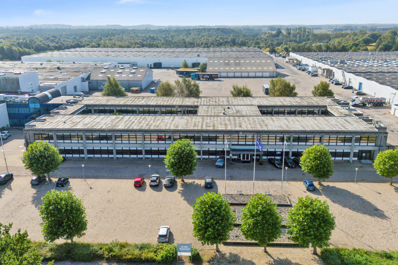 En stor industribygning med fladt tag, flere parkerede biler foran og træer langs indgangen ligger i et stort industriområde.