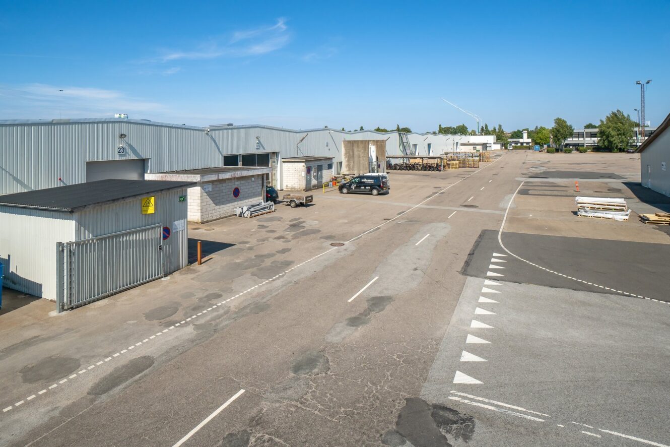 Industriområde med flere lagerbygninger, parkerede biler og stabler af materialer på en bred asfalteret plads under en klar blå himmel.