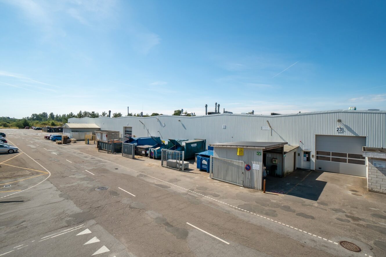 Industriel lagerbygning udvendigt med flere læsseramper, containere og genbrugsbeholdere opstillet langs bygningen under en klar blå himmel.