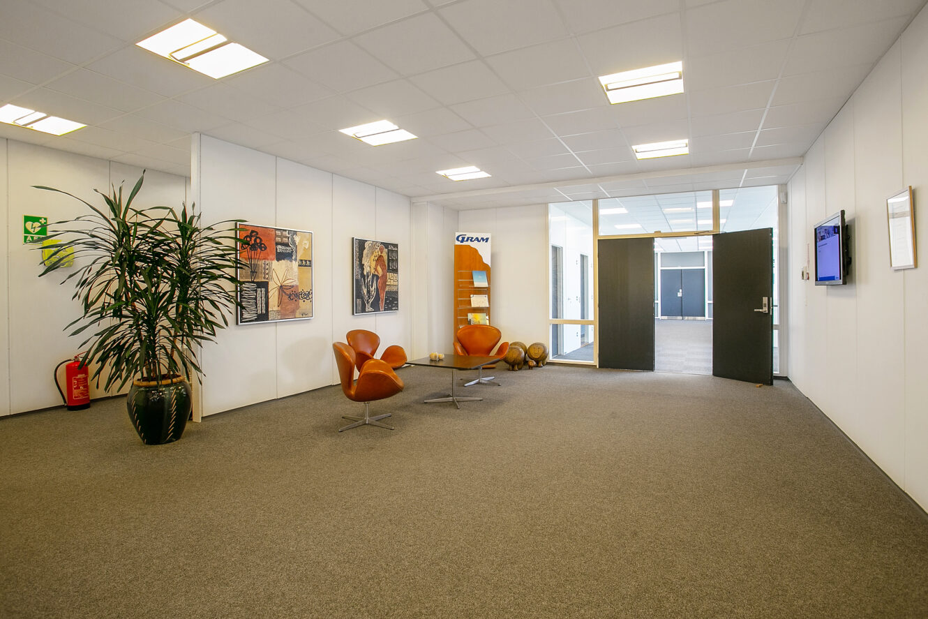 Moderne kontorlobby med gråt tæppe, hvide vægge, to orange stole, en plante, vægkunst, en magasinholder og dobbelte glasdøre, der fører ind til et andet rum.