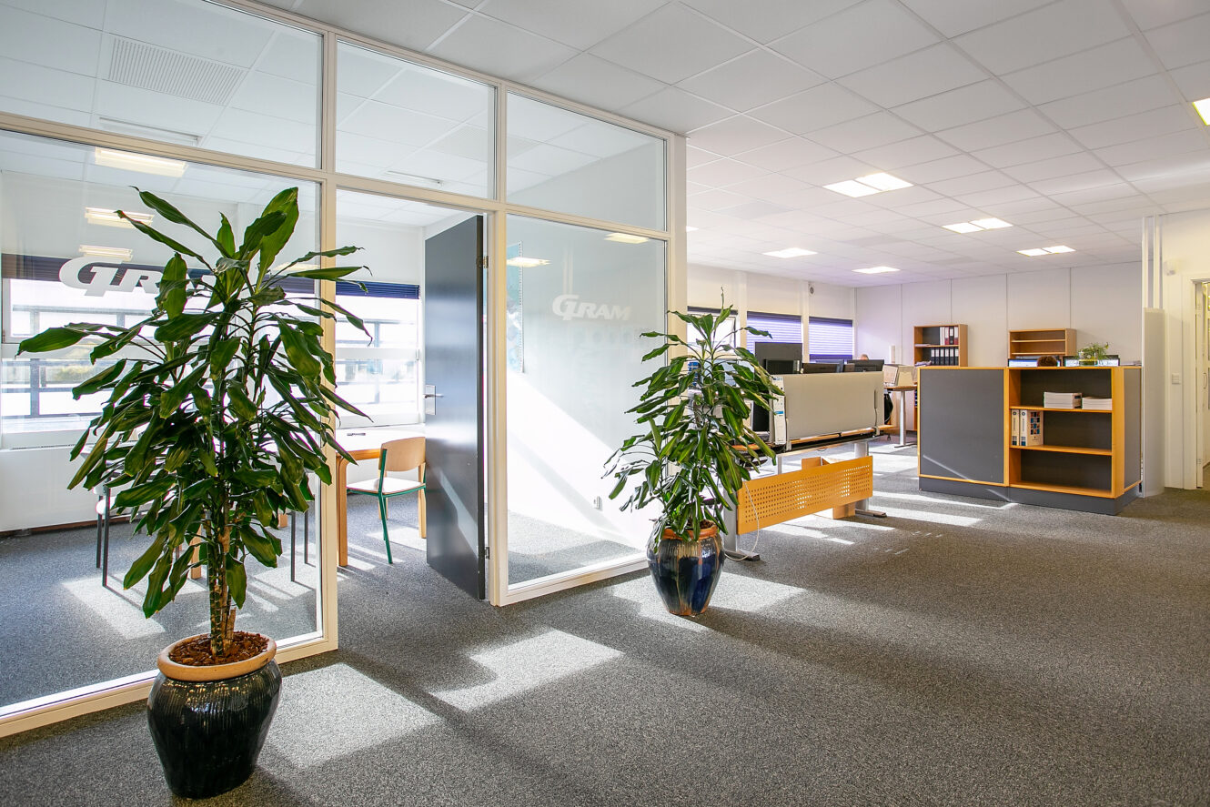 Moderne kontor med mødelokale med glasvægge, potteplanter, skriveborde, hylder og gulvtæppe under lyse loftslamper.