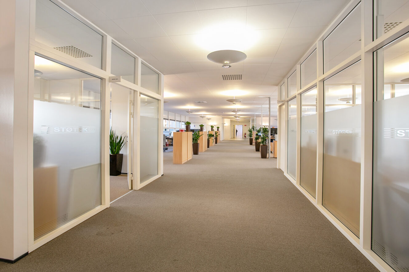 En moderne kontorgang med kontorer med glasvægge på begge sider, lyse tæpper, loftslamper og flere potteplanter langs gangen.