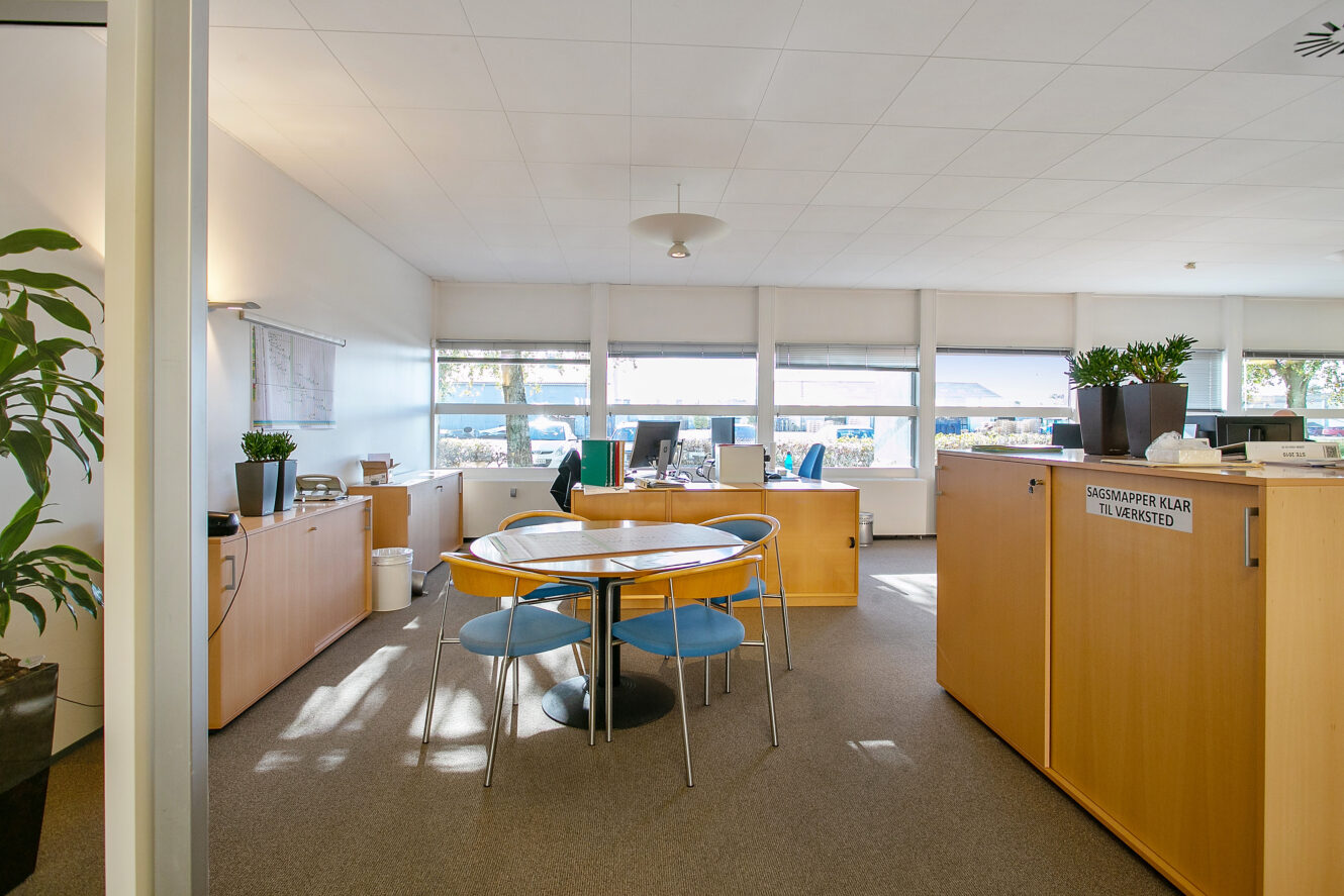 Et lyst kontor med store vinduer, lyse træmøbler, et rundt bord med blå stole og potteplanter på skabe.