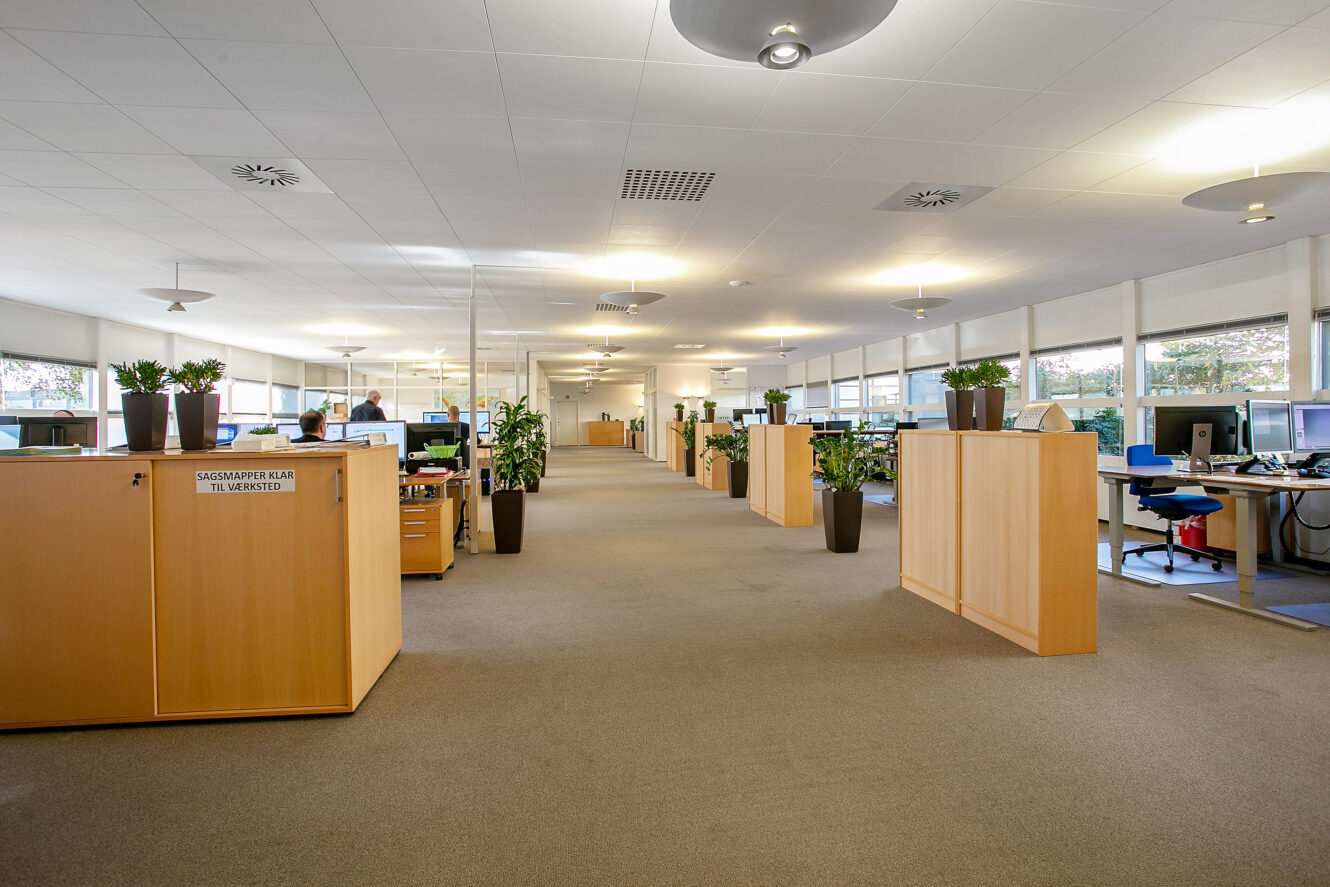 Et rummeligt, moderne åbent kontor med skriveborde, computere, store vinduer, indendørs planter og opbevaringsenheder af træ. Flere mennesker arbejder ved deres skriveborde.