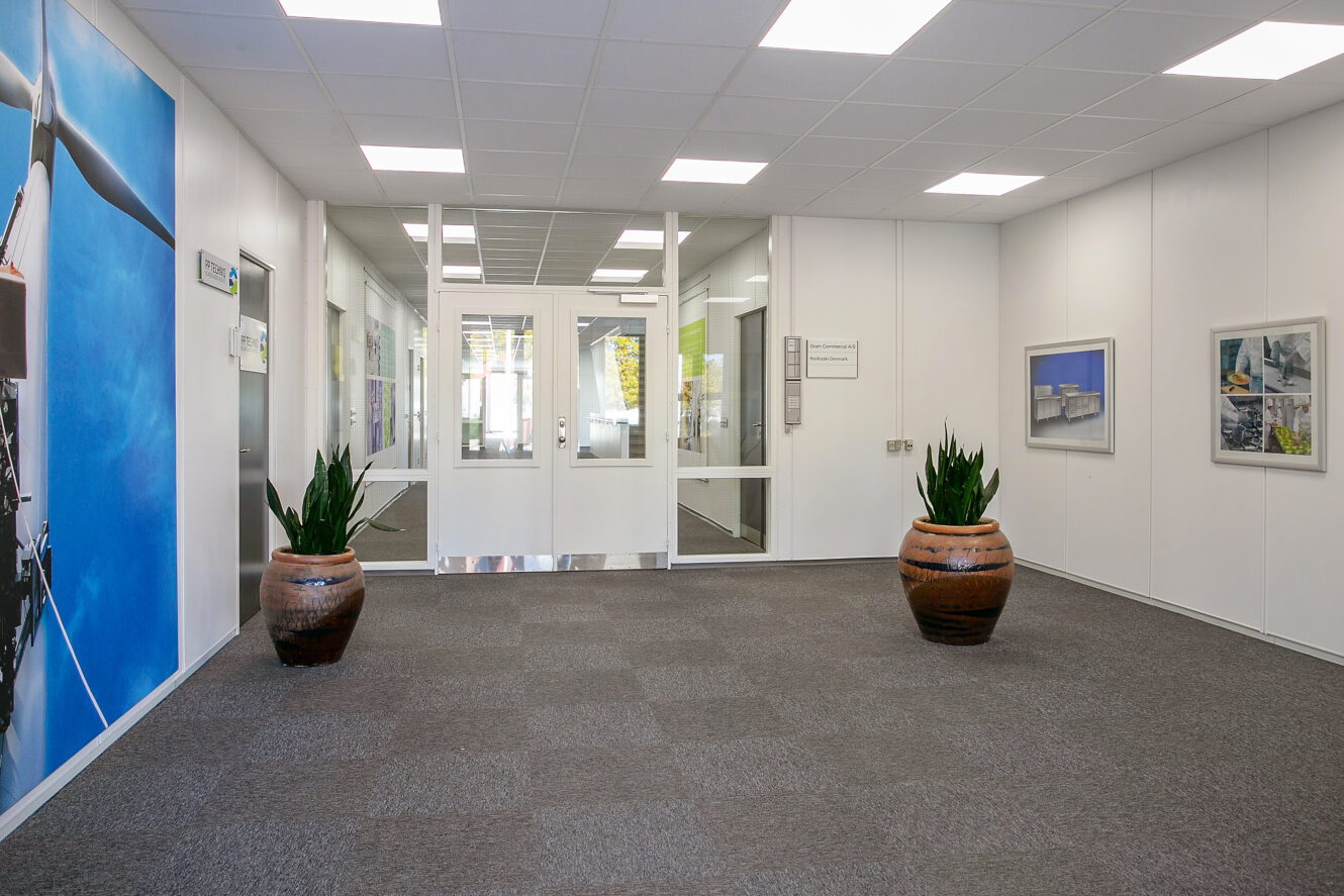 En ren kontorgang med gråt gulvtæppe, hvide vægge, indrammede billeder, to store potteplanter og dobbeltdøre af glas for enden.
