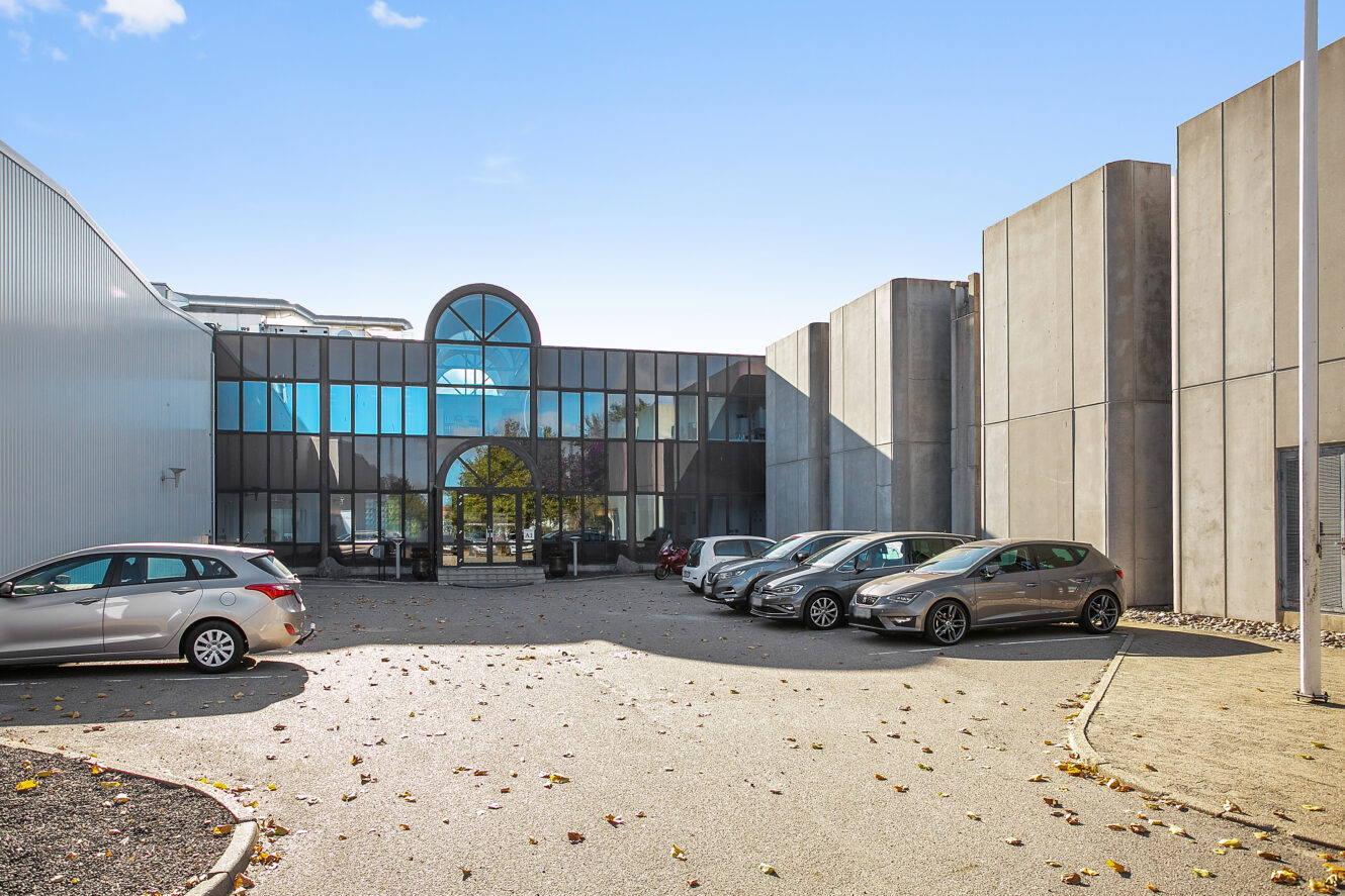 En moderne kontorbygning med store glasvinduer og betonvægge, flere parkerede biler foran og spredte blade på jorden under en klar blå himmel.