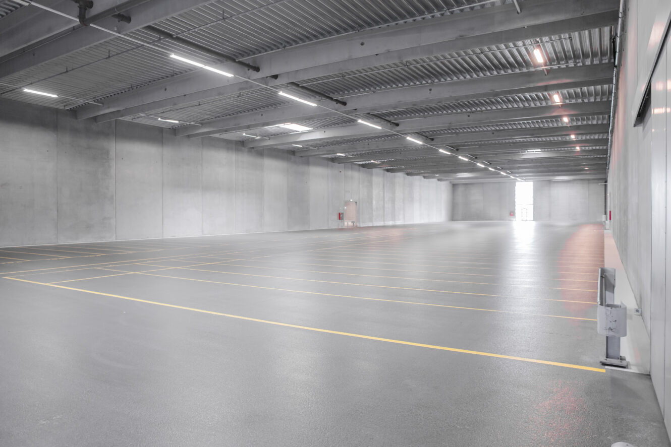 Stort tomt indendørs parkeringshus med grå betongulve, gule parkeringslinjer og synlige loftsbjælker under skarpe lysstofrør.