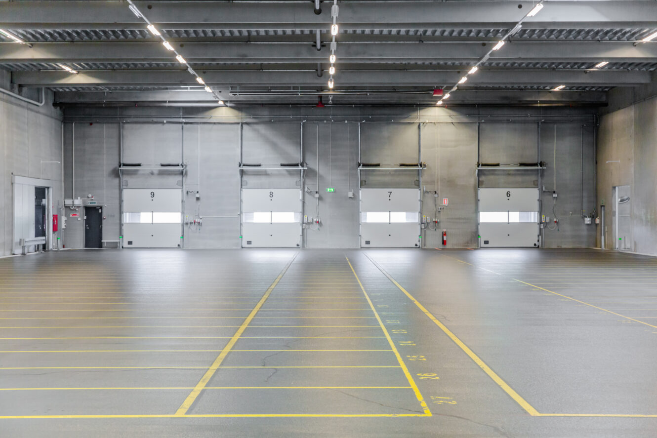 Tomt lager med fem læsseramper, ovenlys og afmærkede parkeringspladser på gulvet.