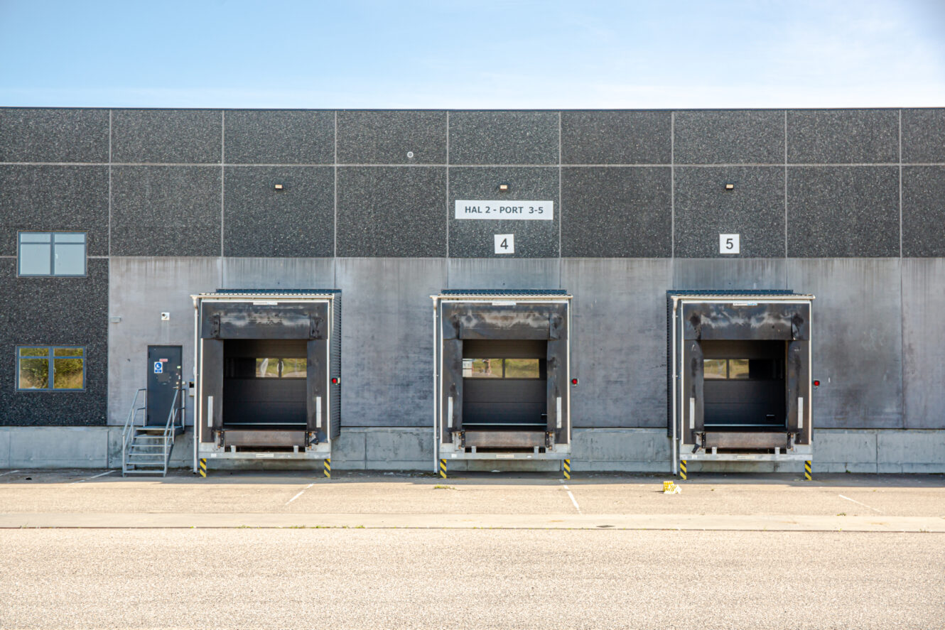 Tre lukkede læsseramper med overliggende baldakiner er nummereret 4, 5 og en umærket midte på en grå industriel lagerbygning.