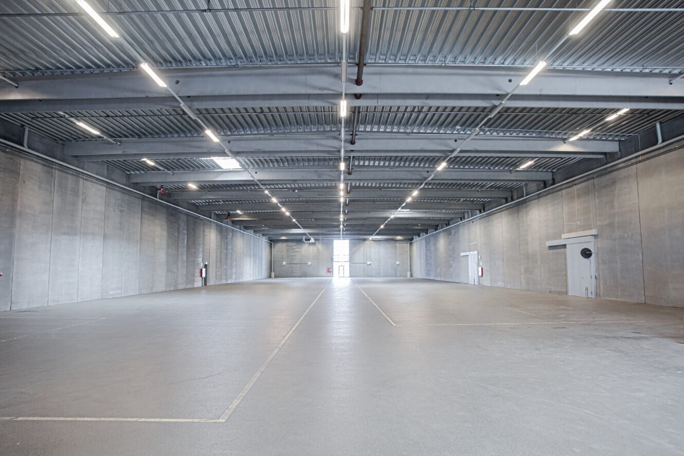 Stort, tomt industrilager med betonvægge, højt til loftet og kraftig ovenlys. Brandslukkere og udgangsdøre er synlige langs siderne.