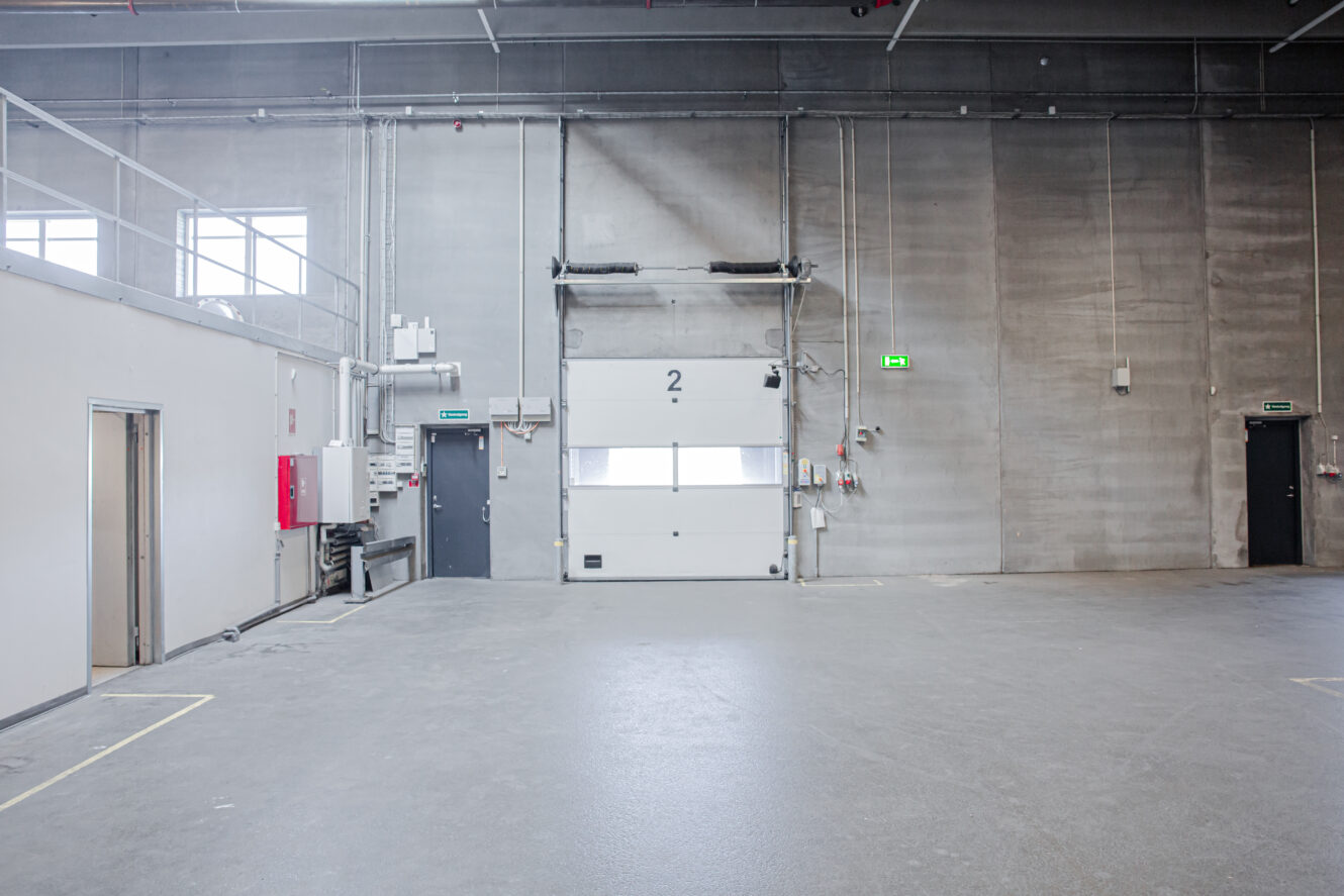 Et tomt industrilager med betongulv, en stor hvid garageport mærket 2 og flere adgangsdøre på venstre og højre væg.