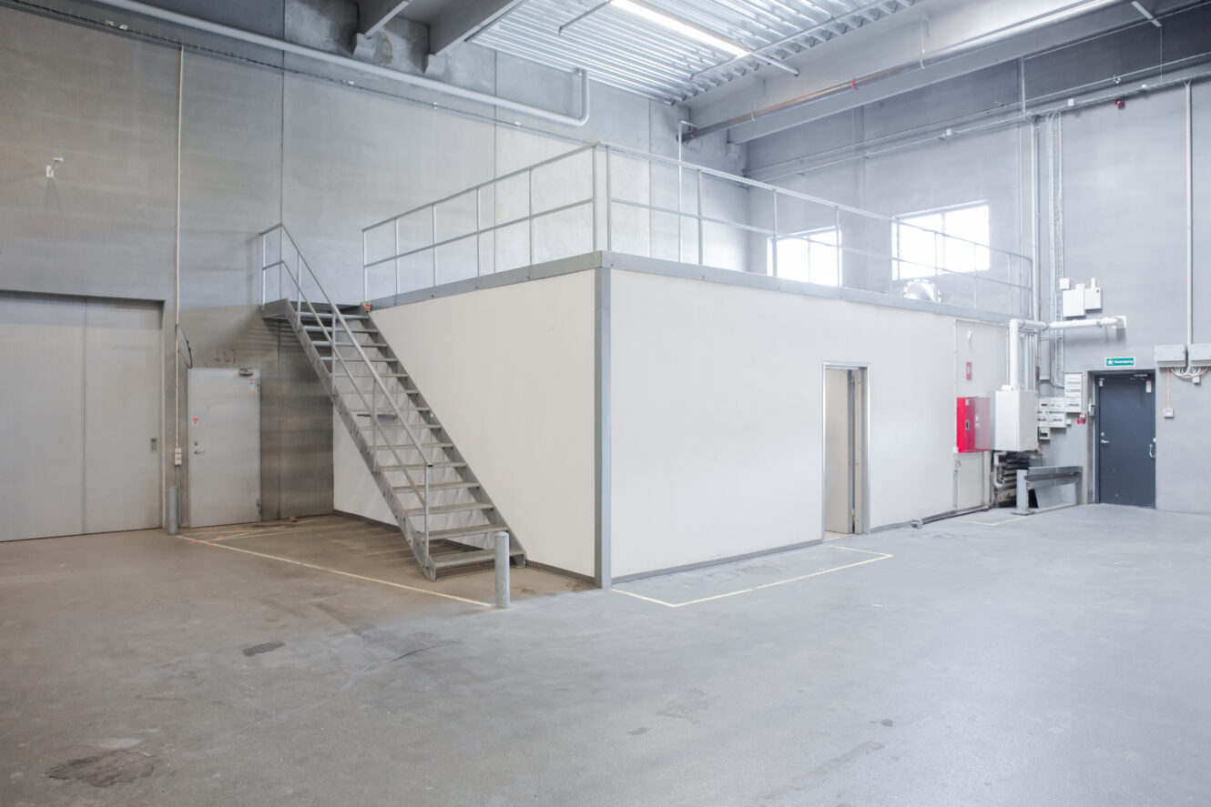 Industrielt lagerinteriør med et hvidt lukket mezzaninkontor, metaltrapper, betongulv og synlige rør og belysning.