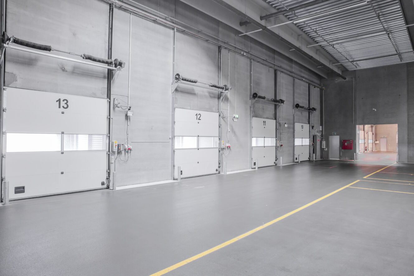 Stor, tom lagerbygning med fire nummererede læsseramper, grå betonvægge og højt til loftet med synlige rør.