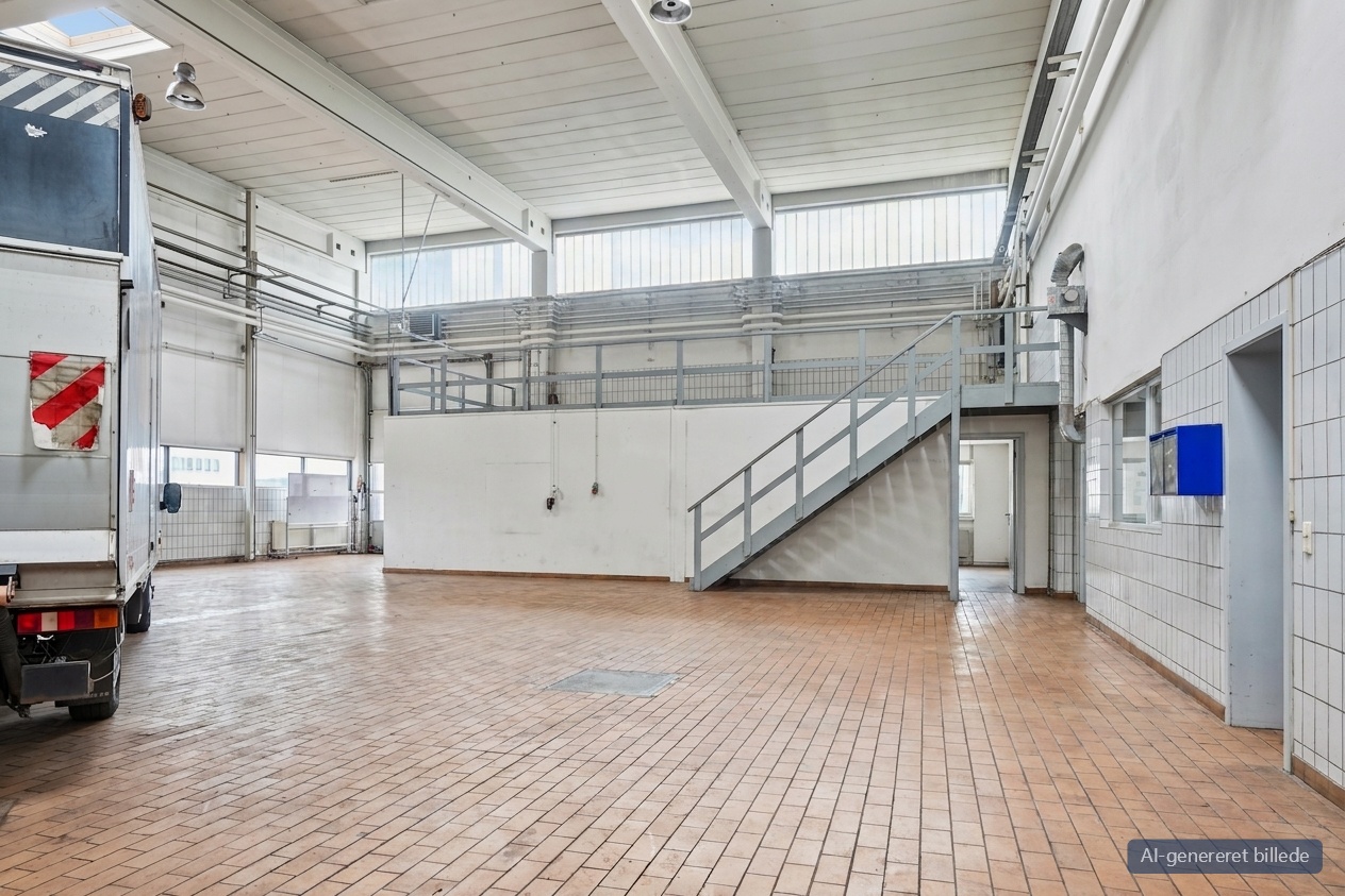 Rummeligt industrielt interiør med klinkegulv, højt til loftet, en delvist synlig lastbil, metaltrapper til en mezzanin og store vinduer.