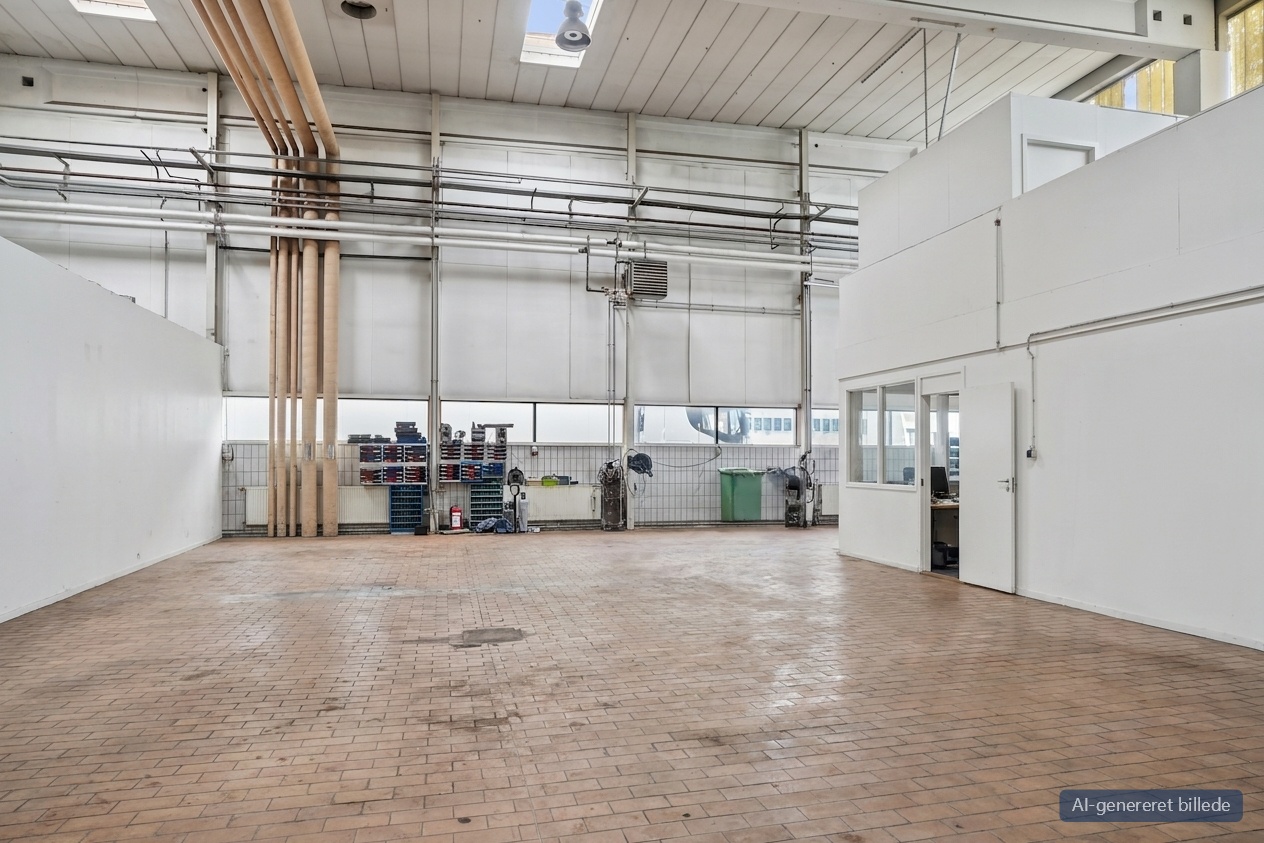 Rummeligt industrielt værksted med klinkegulv, højt til loftet, store vinduer, synlige rør, arbejdsborde og en hvid kontorstruktur.