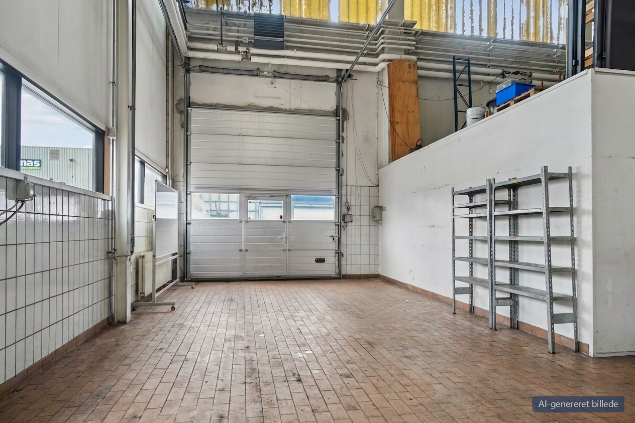 Rummelig industrigarage med flisebelagte vægge, højt til loftet, stor hoveddør og metalhylder. Naturligt lys fra de øverste vinduer.