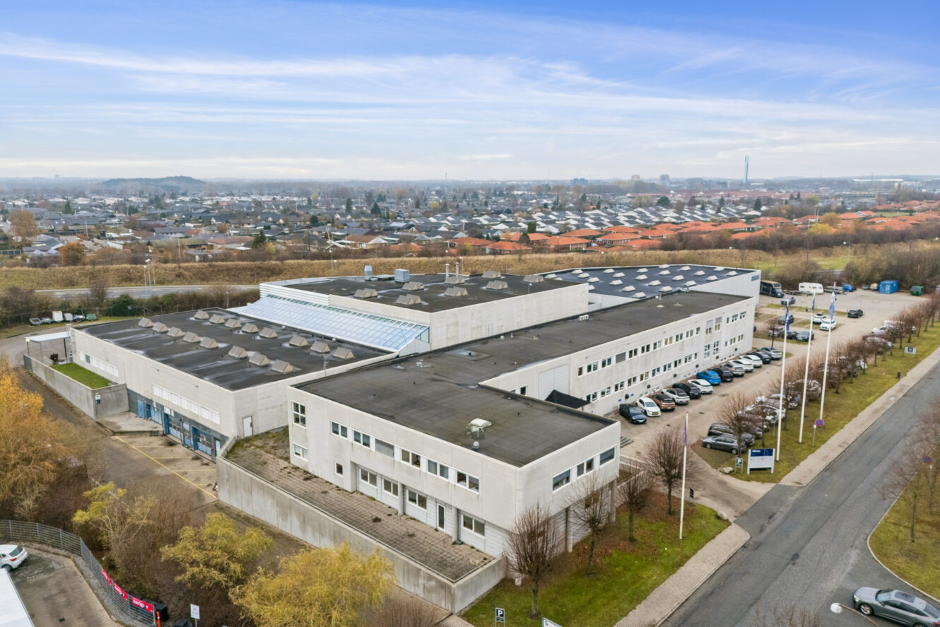 En stor, rektangulær industri- eller kontorbygning med fladt tag og parkeringsplads, omgivet af træer i et forstadsområde under en delvis overskyet himmel.