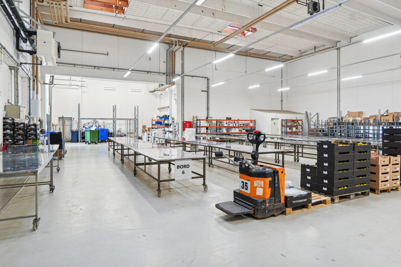 Et rummeligt, lyst industrilager med metalborde, reoler, opbevaringskasser og en lille orange pallevogn mærket »35«.