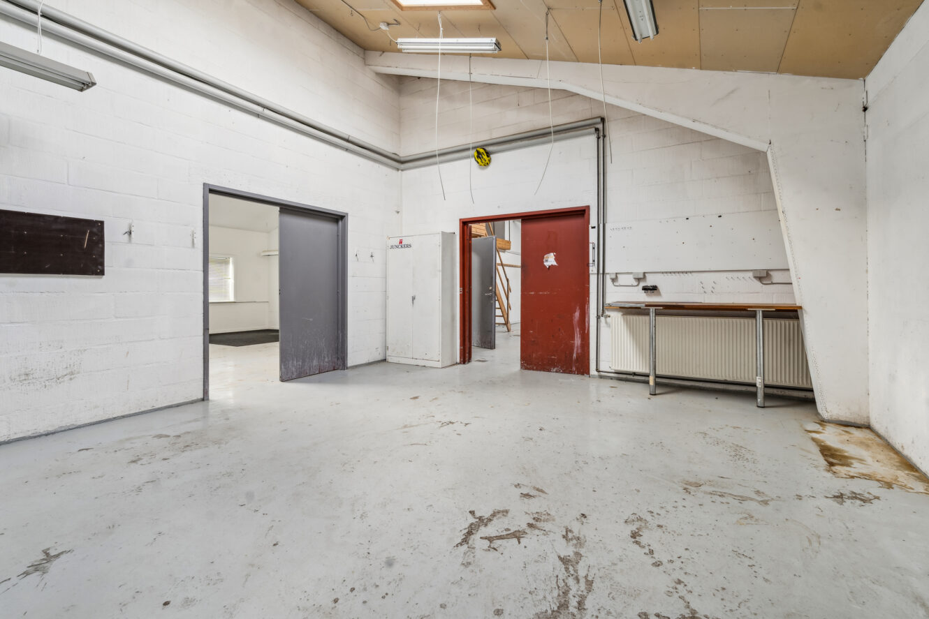 Et tomt industrilokale med hvidmalede murstensvægge, to døråbninger - en grå og en rød - et arbejdsbord, synlige rør og lysstofrør over hovedet.
