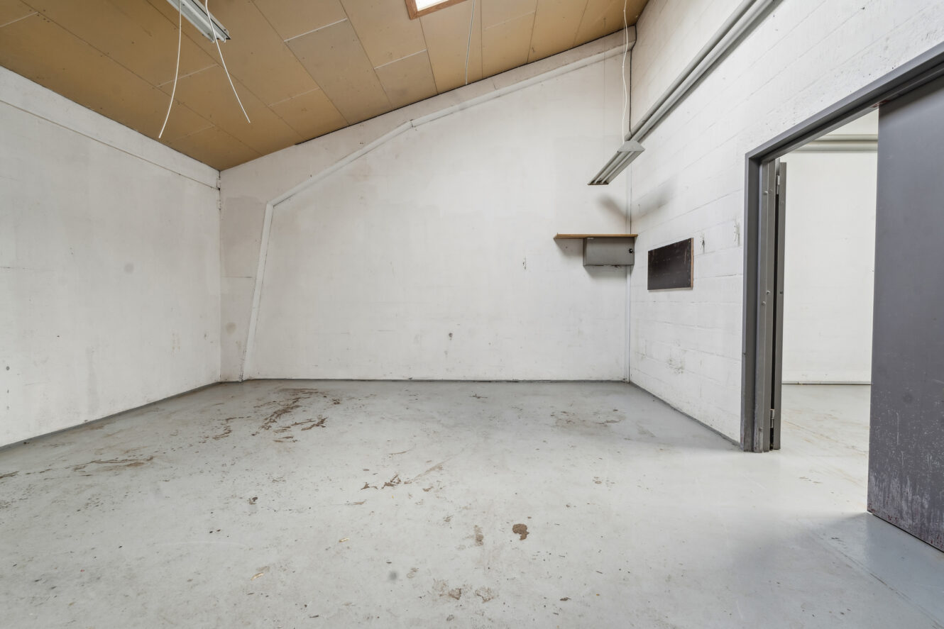 Tomt industrilokale med hvide vægge, betongulv, skråt loft, ovenlys, en lille hylde og en åben dør i højre side.