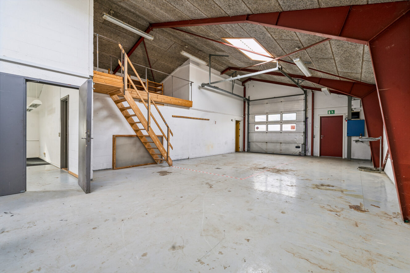 Industrielt lagerinteriør med betongulv, højt til loftet, metalbjælker, en trætrappe, der fører op til en hems, og en stor garageport.