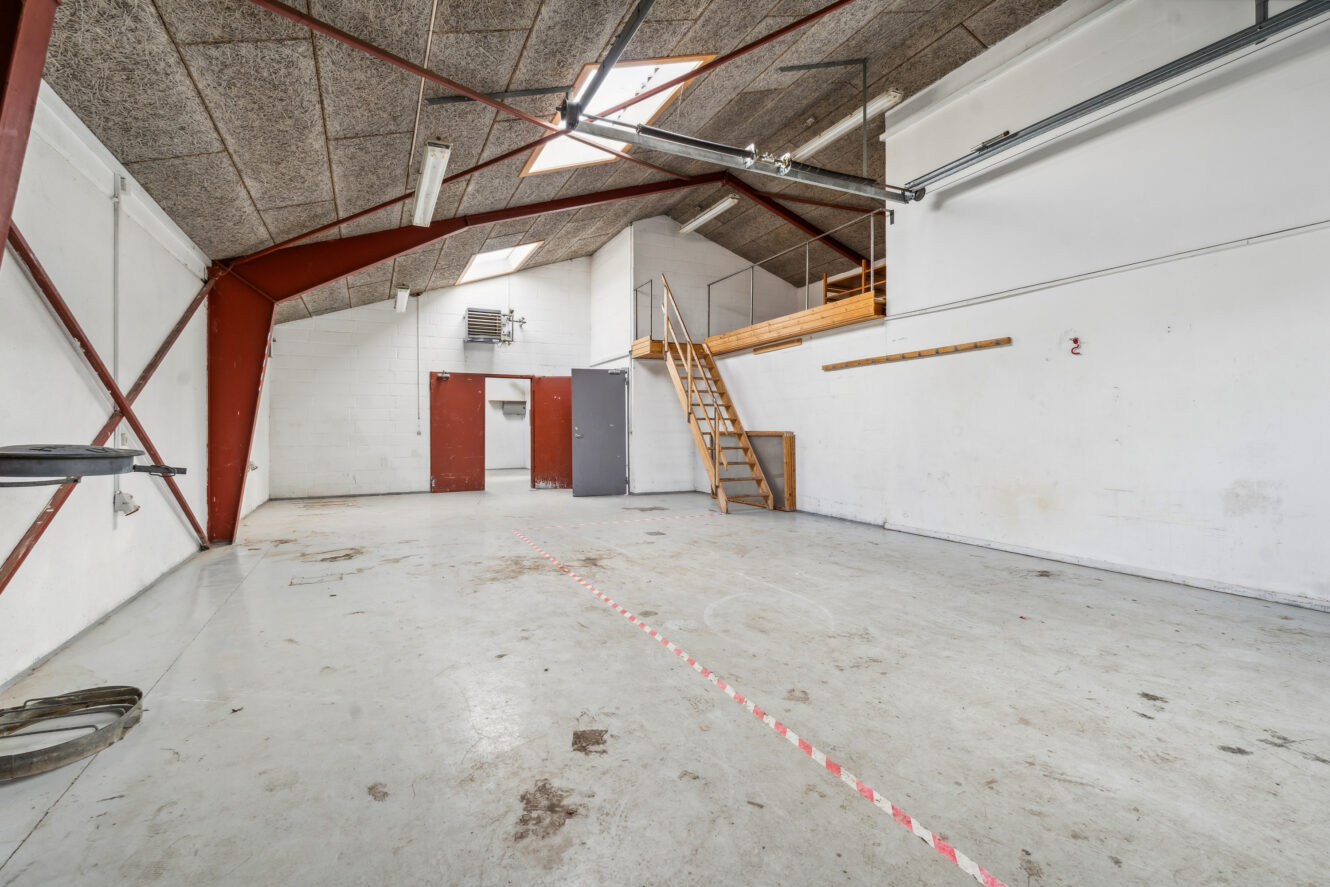 Tomt industrielt lagerinteriør med betongulv, højt til loftet, ovenlys, røde stålbjælker og en træloft med adgang via trapper.