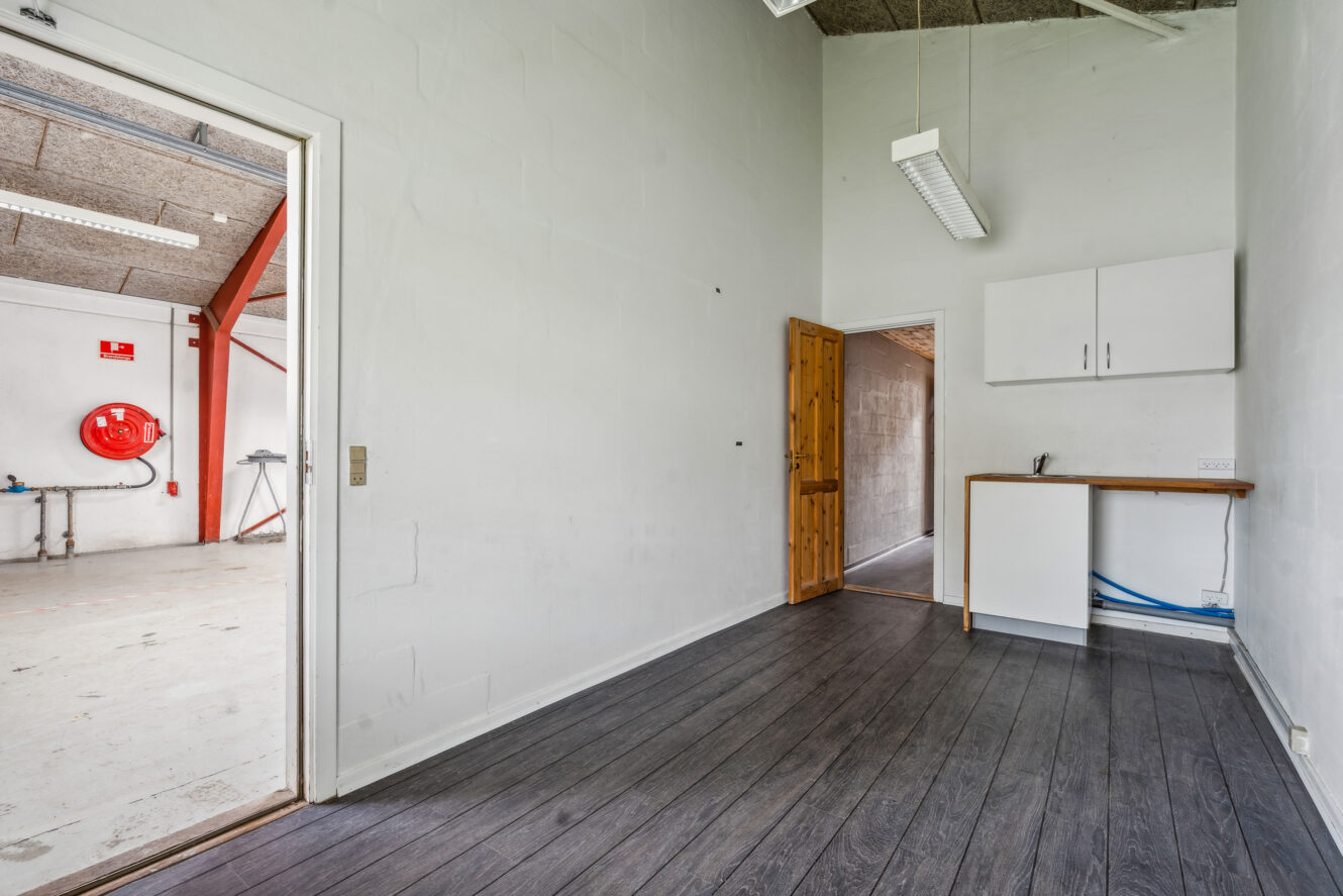 Et lille tomt rum med mørkt trægulv, en trædør, hvide vægge, et lysarmatur, overskabe og et tekøkken; et tilstødende industriområde er synligt gennem en åben døråbning.