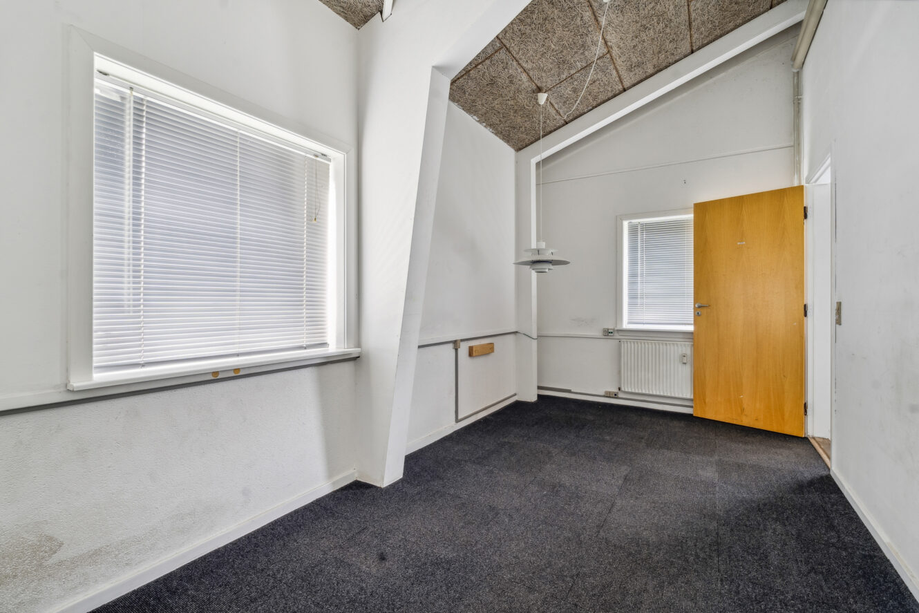 Tomt rum med mørkt gulvtæppe, hvide vægge, to vinduer med persienner, en radiator, en loftslampe og en åben trædør.