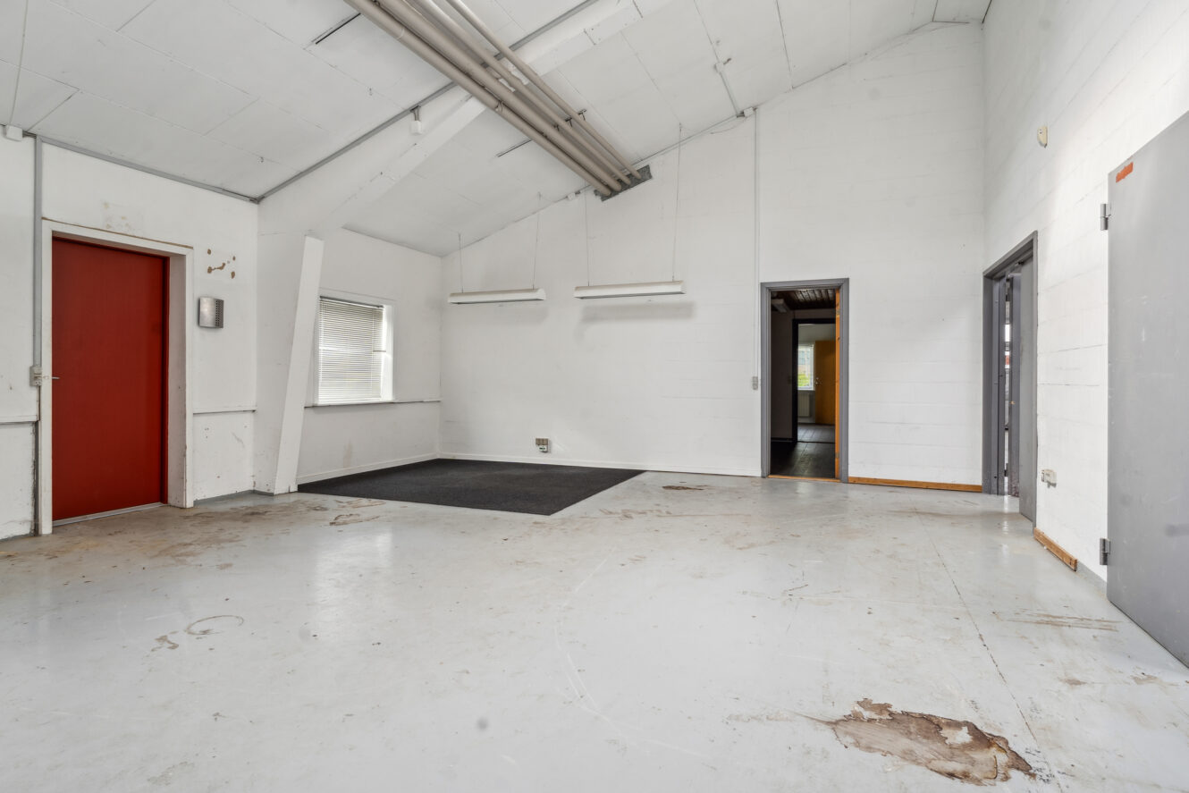 Tomt industrilokale med hvidmalede vægge, synlige rør i loftet, store vinduer, to åbne døråbninger, en rød dør og en sort dørmåtte på gulvet.