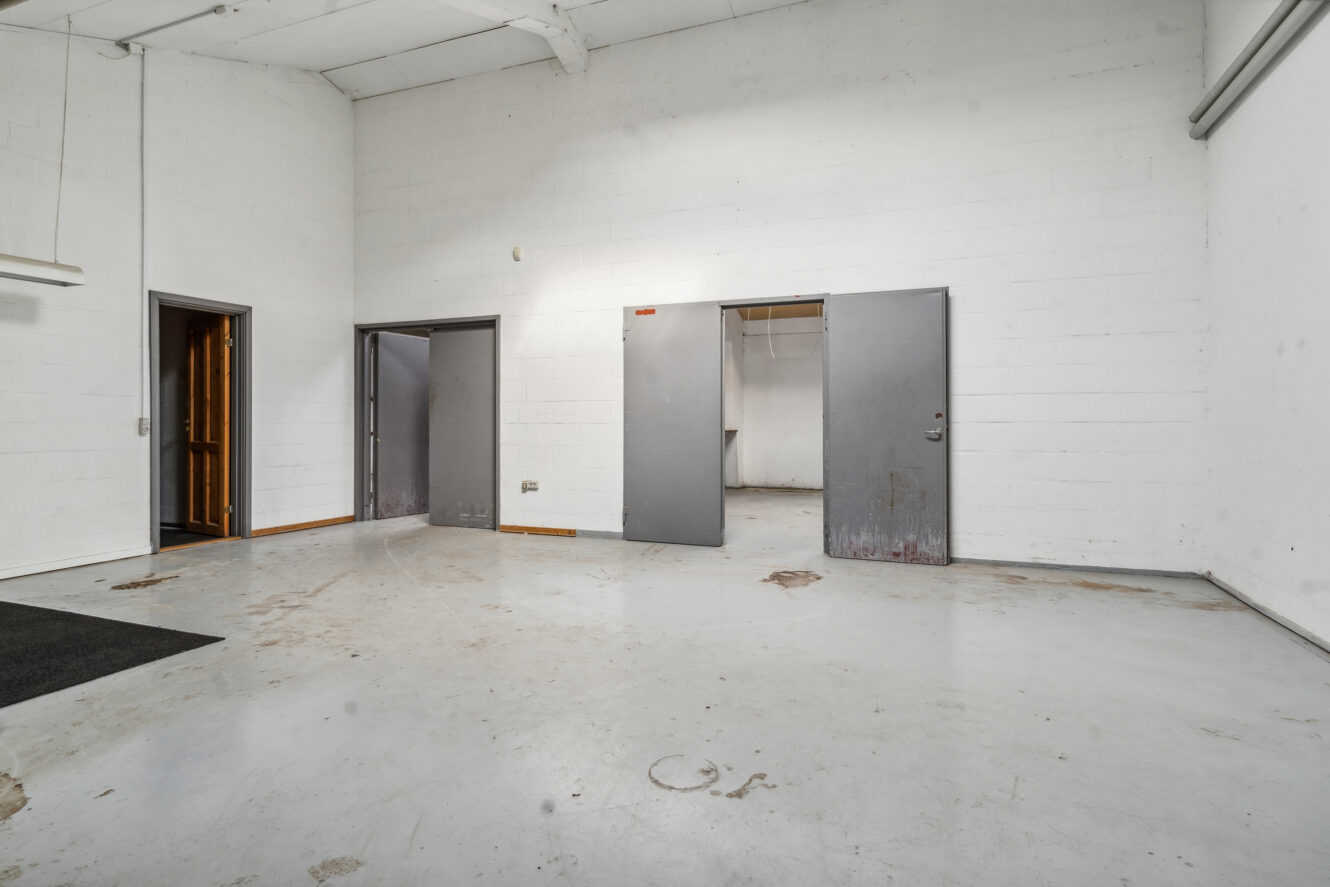 Tomt industrilokale med hvidmalede murstensvægge, betongulv og tre åbne metaldøre, der afslører mindre tilstødende rum.