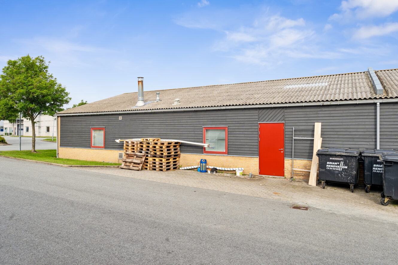 Grå industribygning i én etage med rød dør og vinduesrammer, træpaller stablet udenfor og sorte skraldespande langs siden på en asfalteret gade.