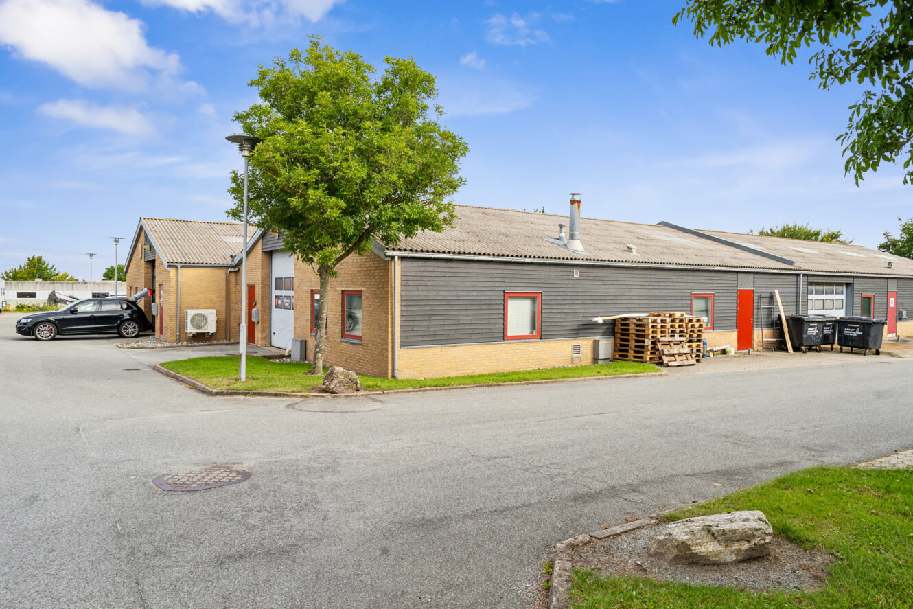 Erhvervsbygning i én etage med grå facadebeklædning, gule murstensundervægge og røde vinduer, omgivet af en parkeringsplads, træer og et par parkerede biler.