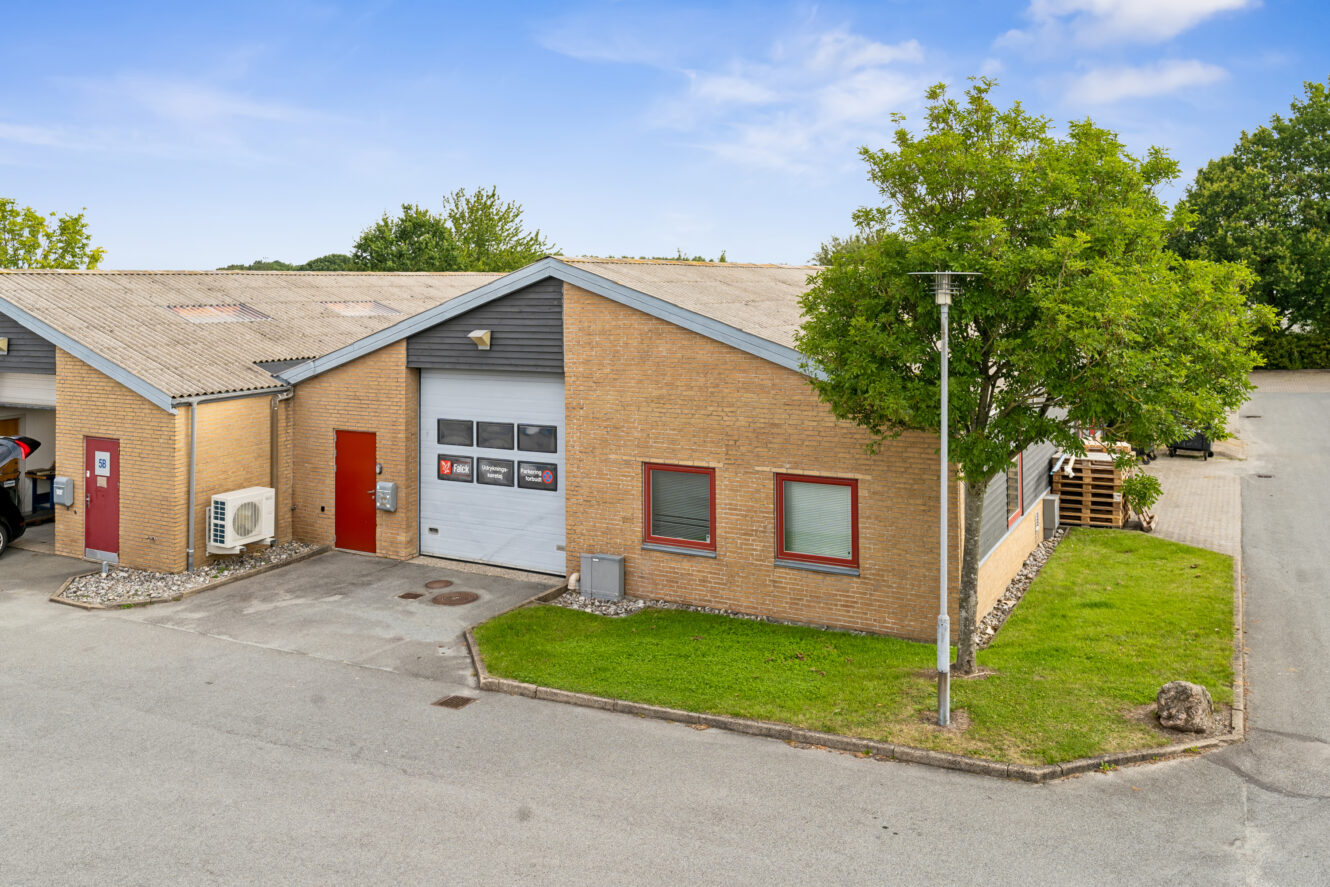 Industribygning i én etage med murstensfacade, røde døre og en garageindgang, omgivet af græs, træer og en asfalteret indkørsel under en blå himmel.