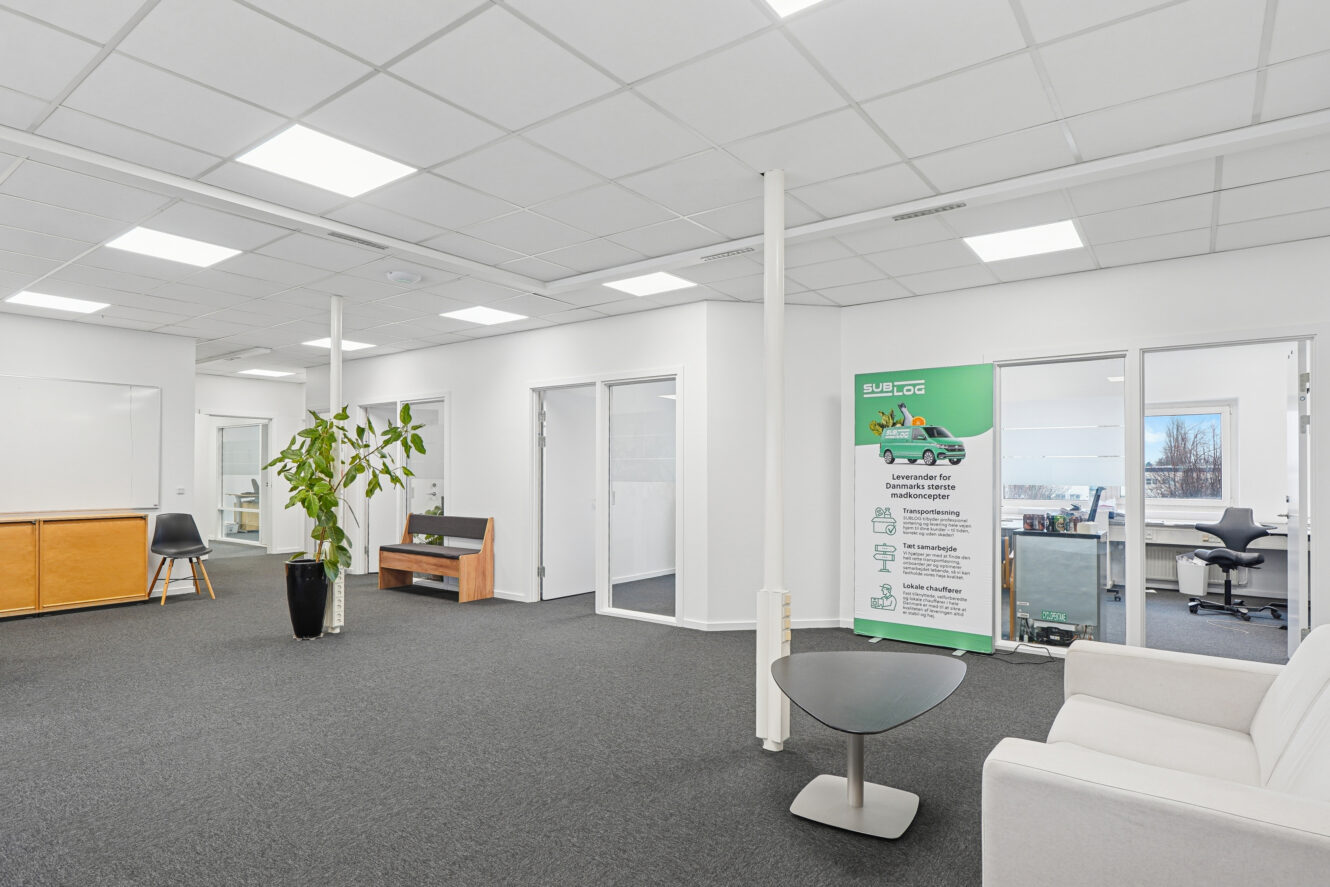 Moderne kontor med hvide vægge, glasskillevægge, en hvid sofa, et lille bord, en potteplante og et reklamebanner ved en dør med vindue.