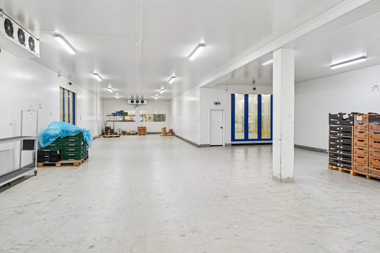 Stort, rent industrilager med hvide vægge og loft, stablede paller, papkasser, ovenlys og udstyr under en blå presenning.