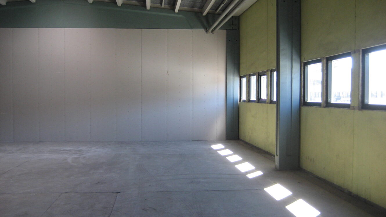 Tomt industrilokale med betongulv, ubehandlede vægge, synlige loftsbjælker og en række rektangulære vinduer, der lukker sollys ind i højre side.