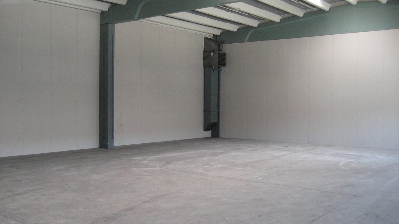 Et tomt industrilokale med betongulv, hvide vægge, synlige loftsbjælker og et væghængt varmeapparat i hjørnet.