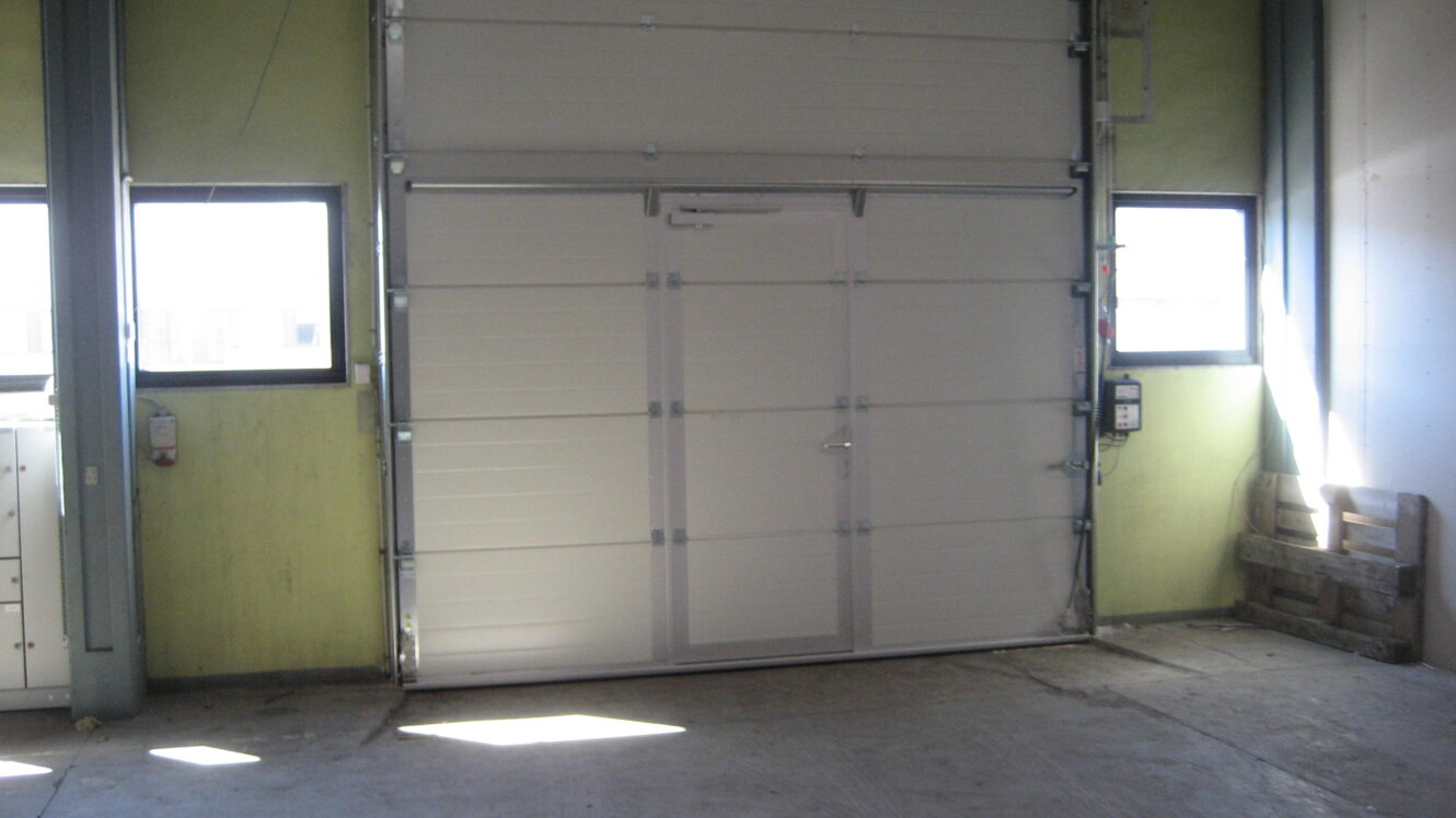 En stor hvid industriel garageport med en mindre fodgængerdør indeni, flankeret af to vinduer i en lagerbygning.