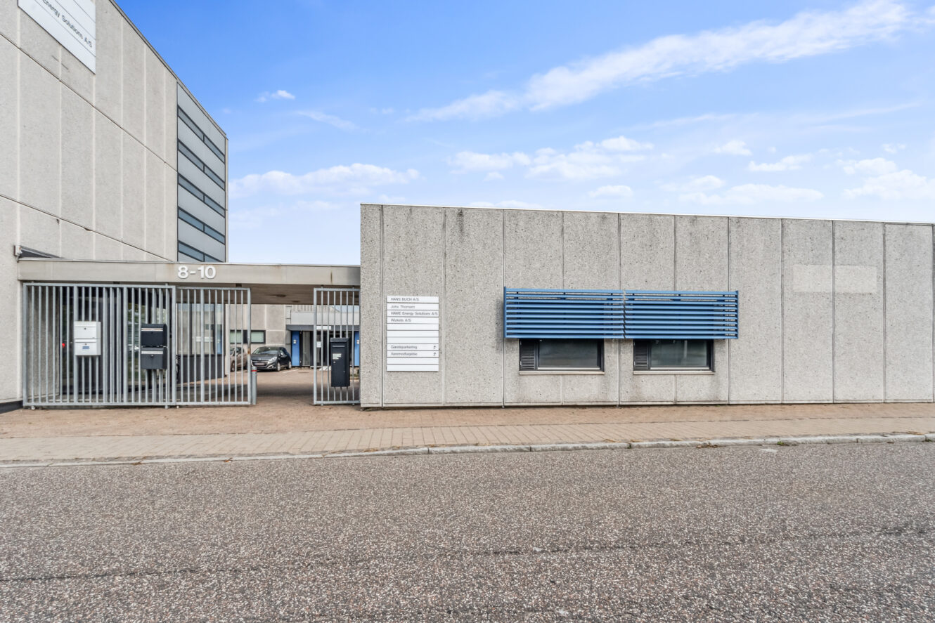 En moderne industribygning med en indhegnet indgang, grå ydervægge, blå vinduesmarkise og skiltning nær indgangen under en delvis overskyet himmel.