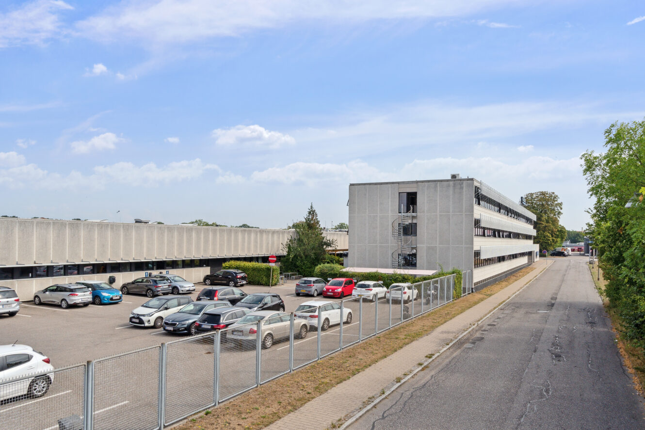 En parkeringsplads med flere parkerede biler ved siden af en grå kontorbygning i flere etager og et indhegnet fortov under en delvis overskyet himmel.