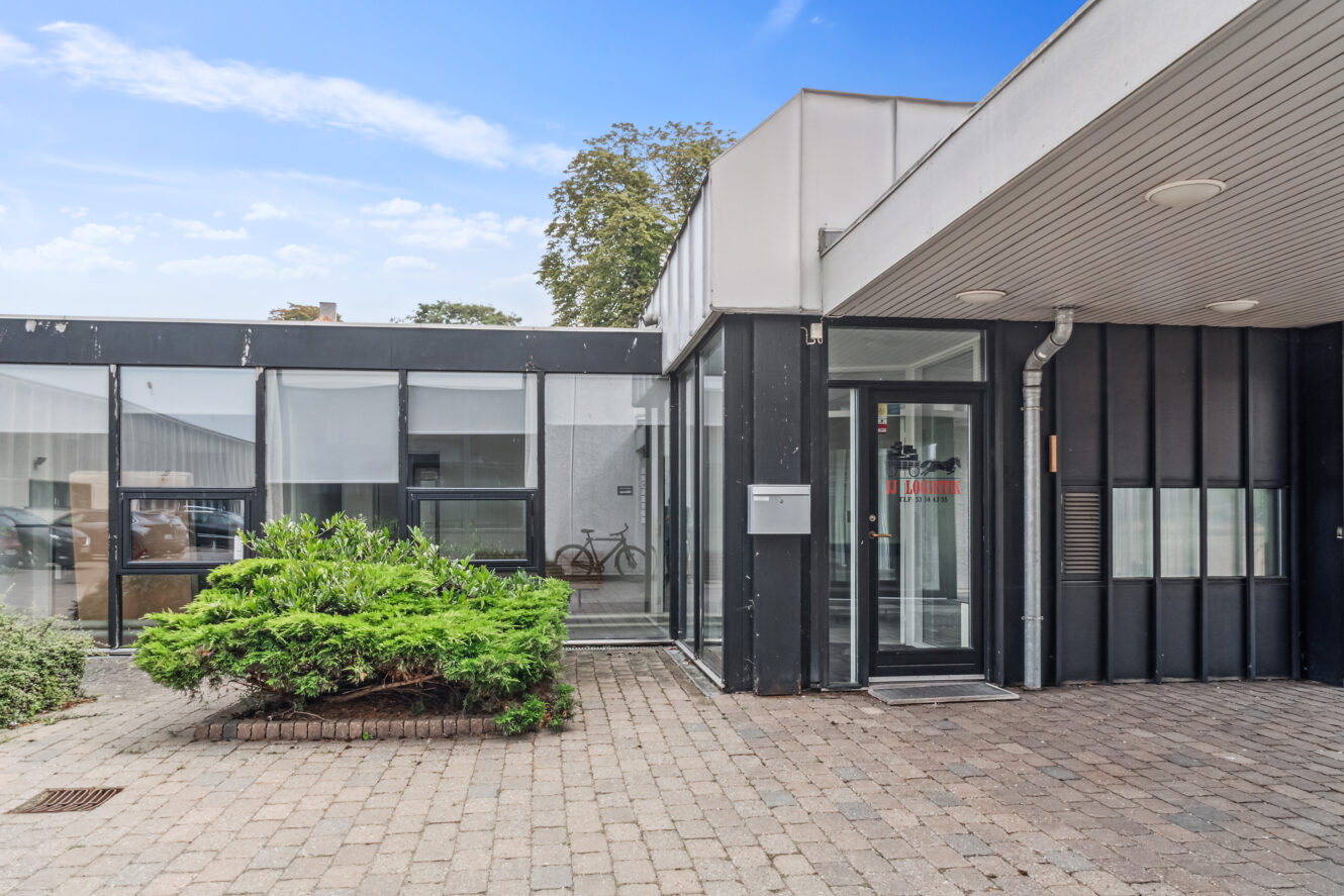 Moderne bygning med store vinduer, indgangsdør i glas, udhæng og murstensbelagt gangbro. En cykel er synlig gennem vinduet.